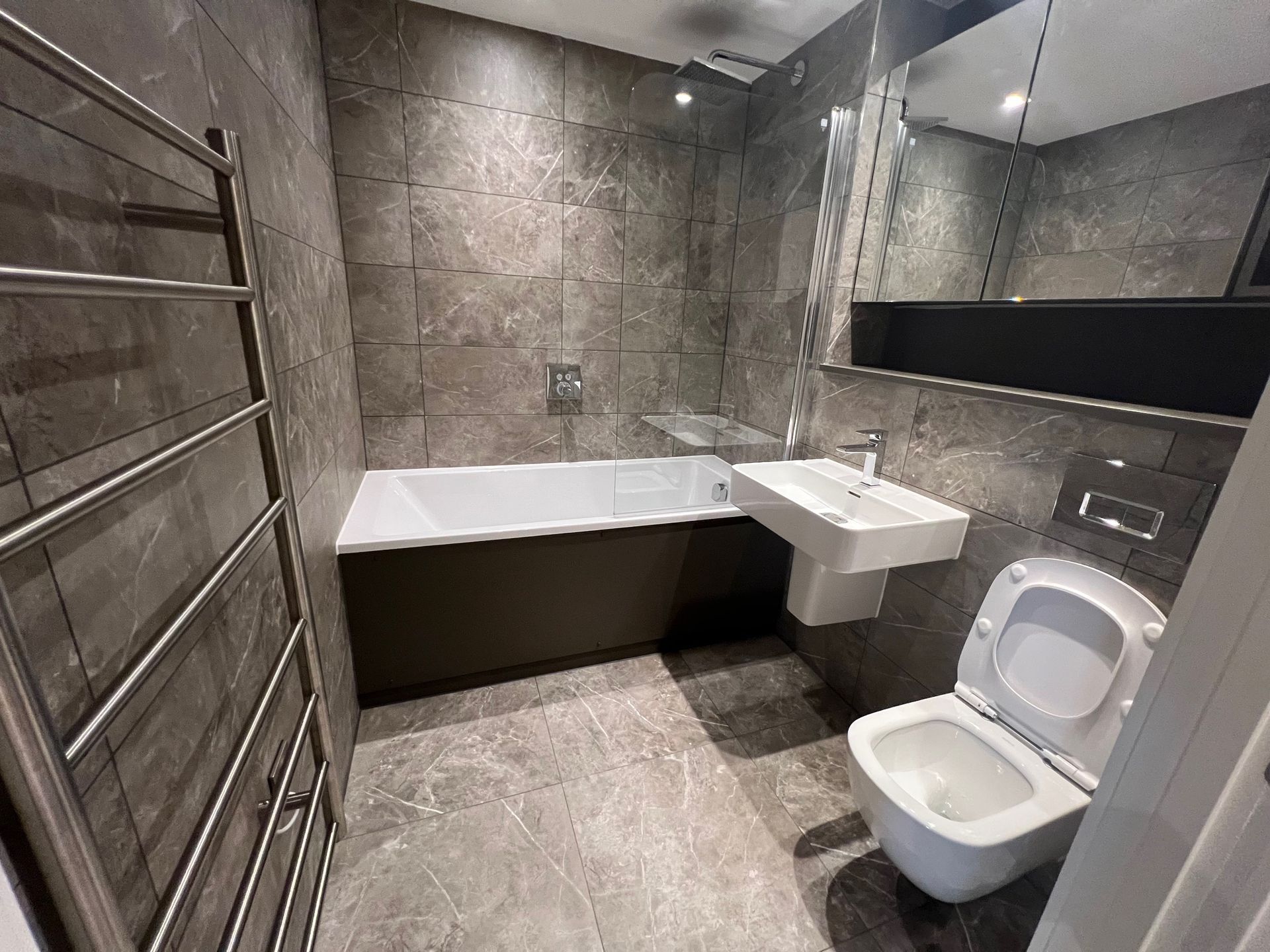 Modern bathroom with gray tile walls and floor, bathtub, toilet, and sink.