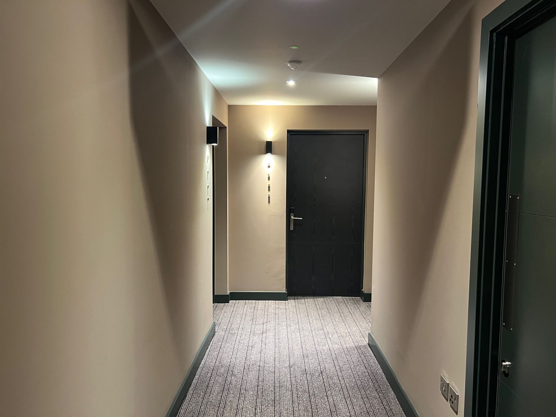 Narrow hotel hallway with a patterned carpet, beige walls, and a black door.