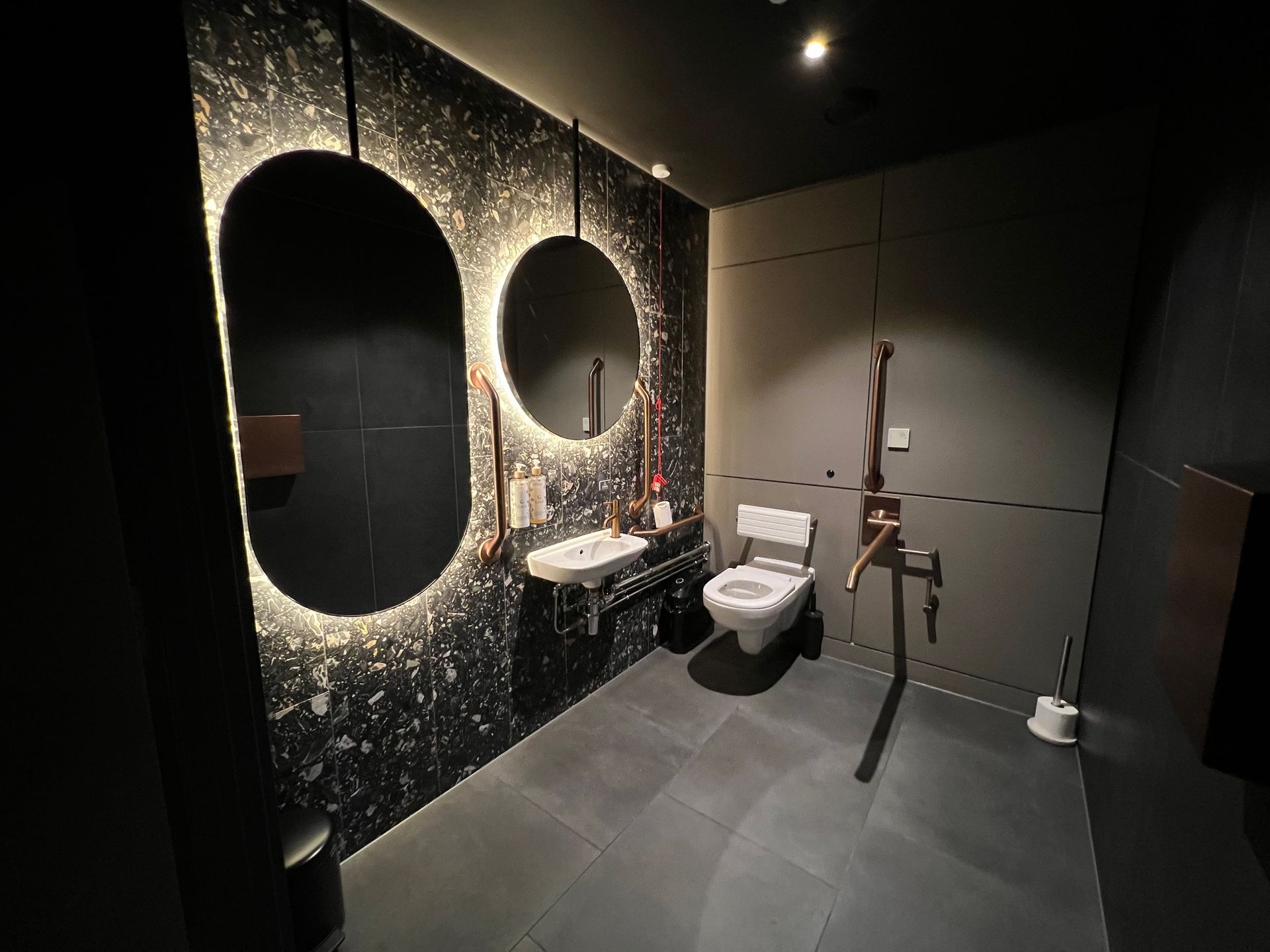 Modern restroom with dark decor, lit mirrors, accessible toilet, and copper fixtures.