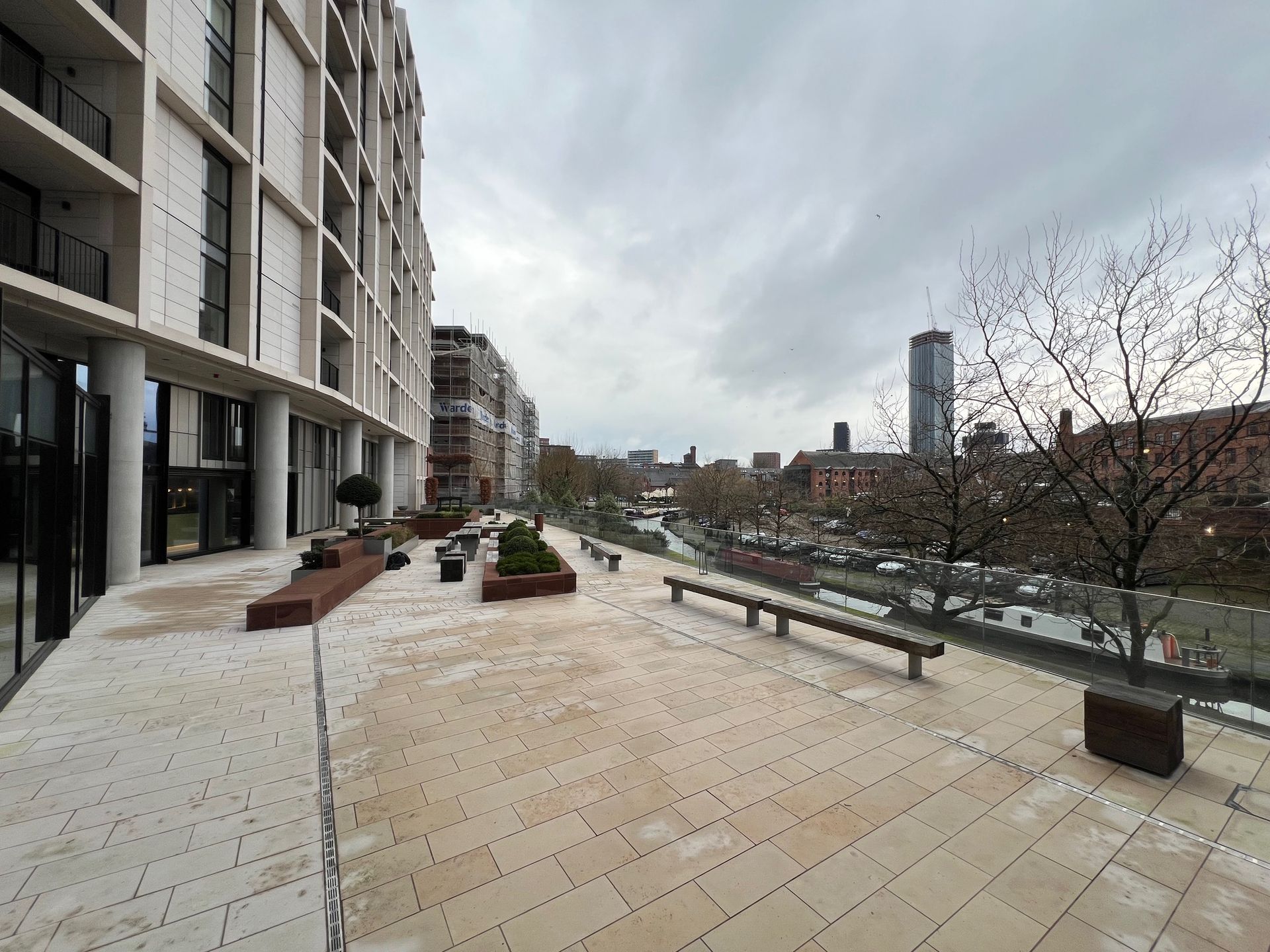 Exterior view of a modern building's patio with benches, planters, and a cityscape in the background under a cloudy sky.