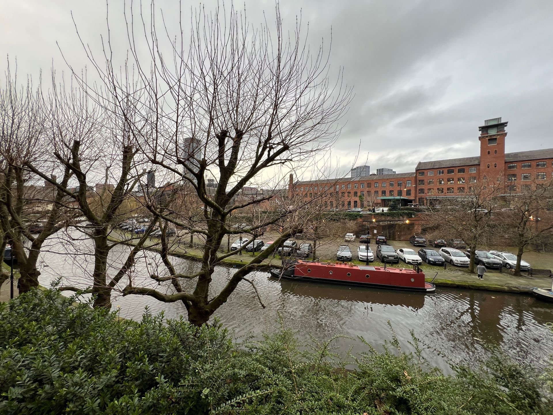 Trees over a canal, red boat, cars, and brick building with a tower under an overcast sky.