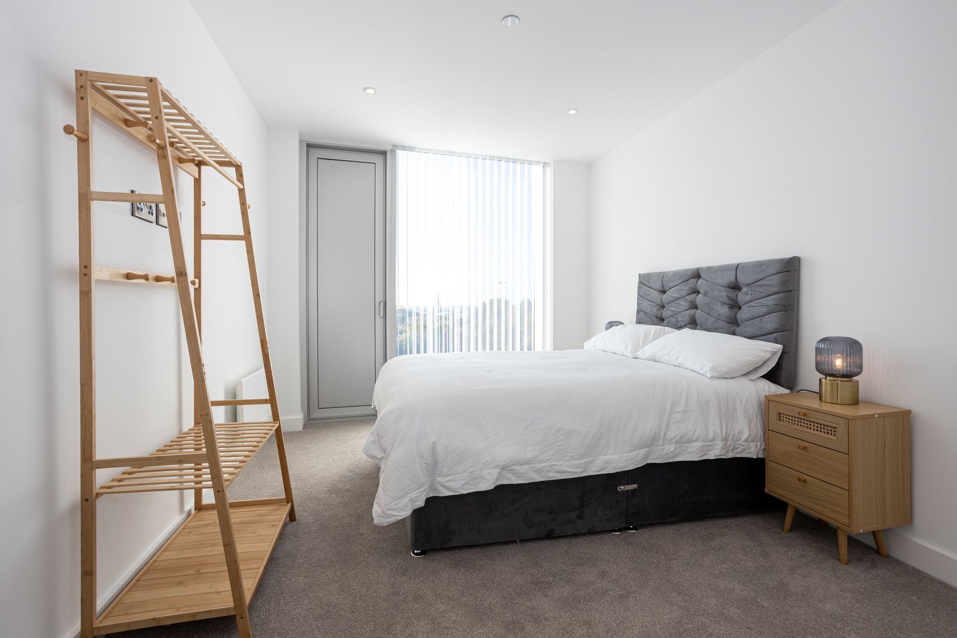 Bedroom with bed, nightstand, and clothing rack. Grey carpet and white walls.