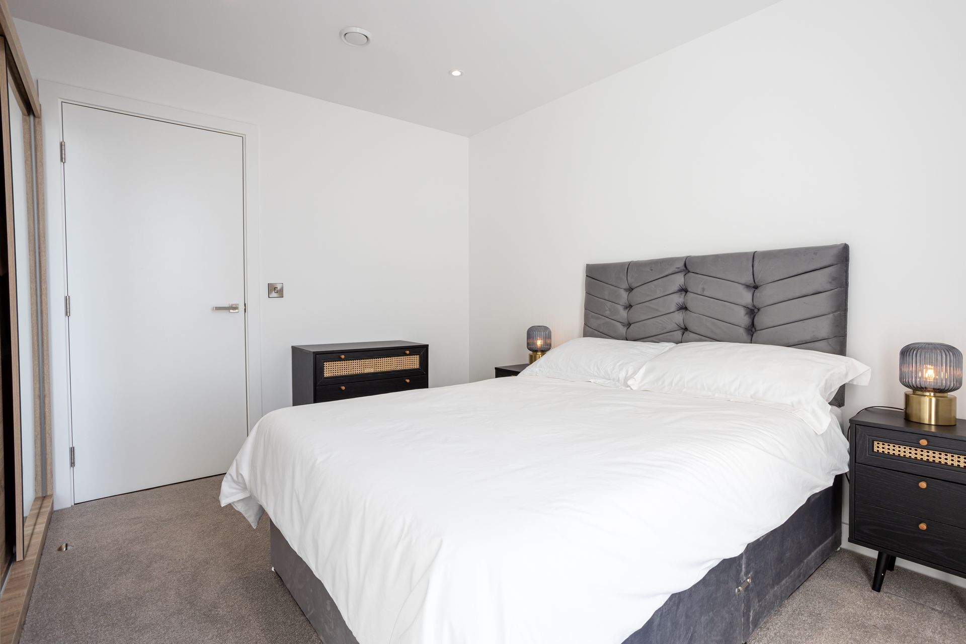 Bedroom with a bed, nightstands, closet, and a closed door. White walls, gray carpet, and dark furniture.