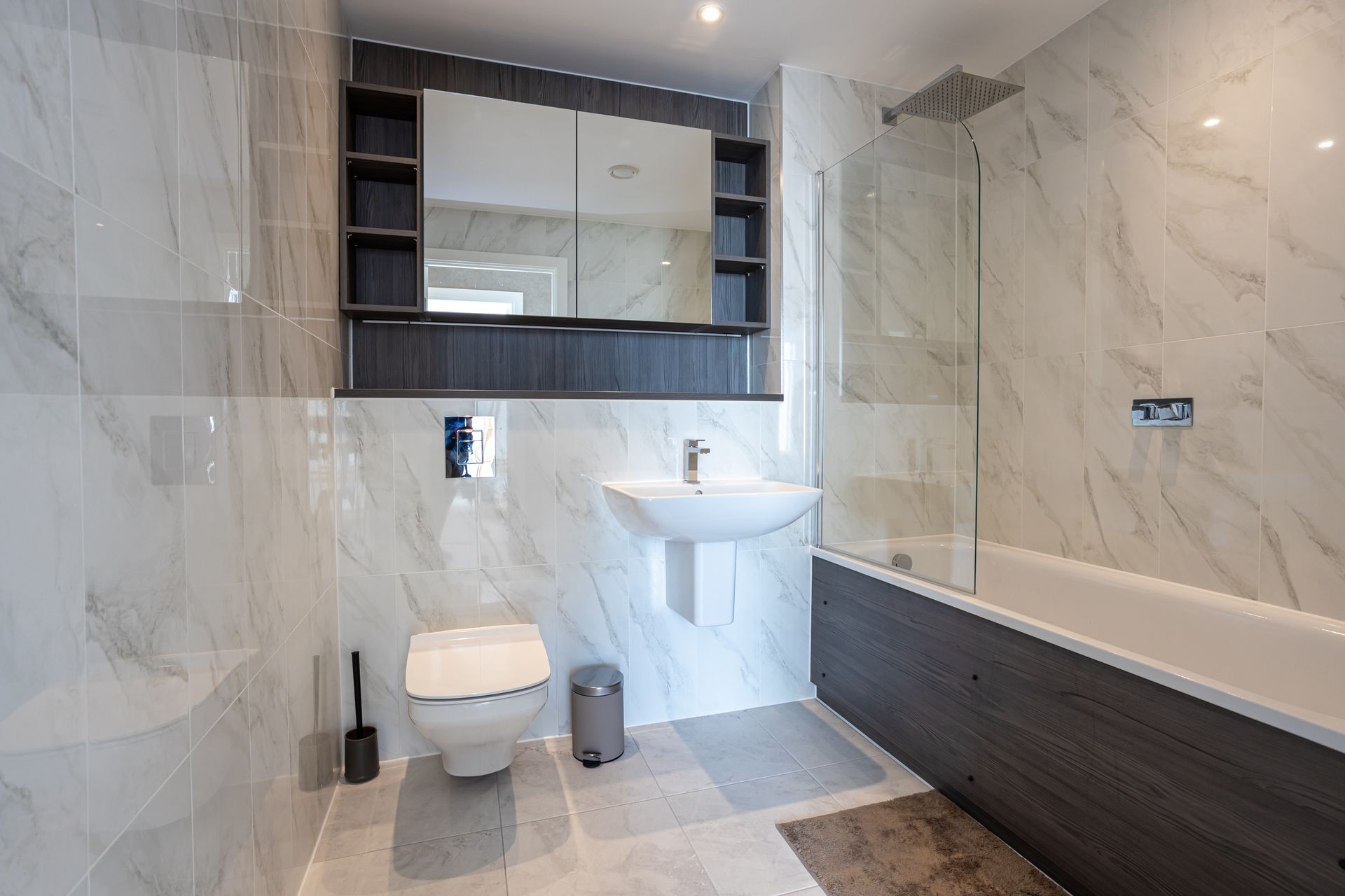 Modern bathroom with marble-look tiles, wall-mounted sink, toilet, dark wood vanity, and a shower/tub combo.