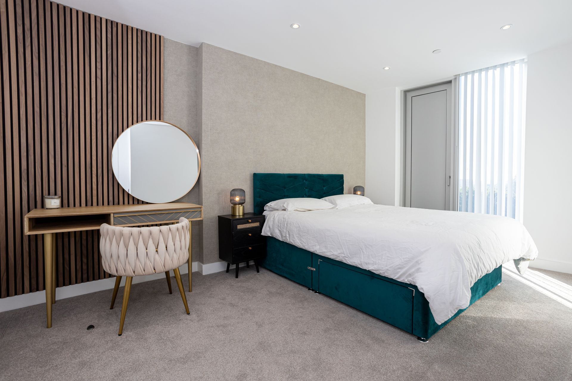 Bedroom with a teal bed, wooden accent wall, vanity, and window with blinds.