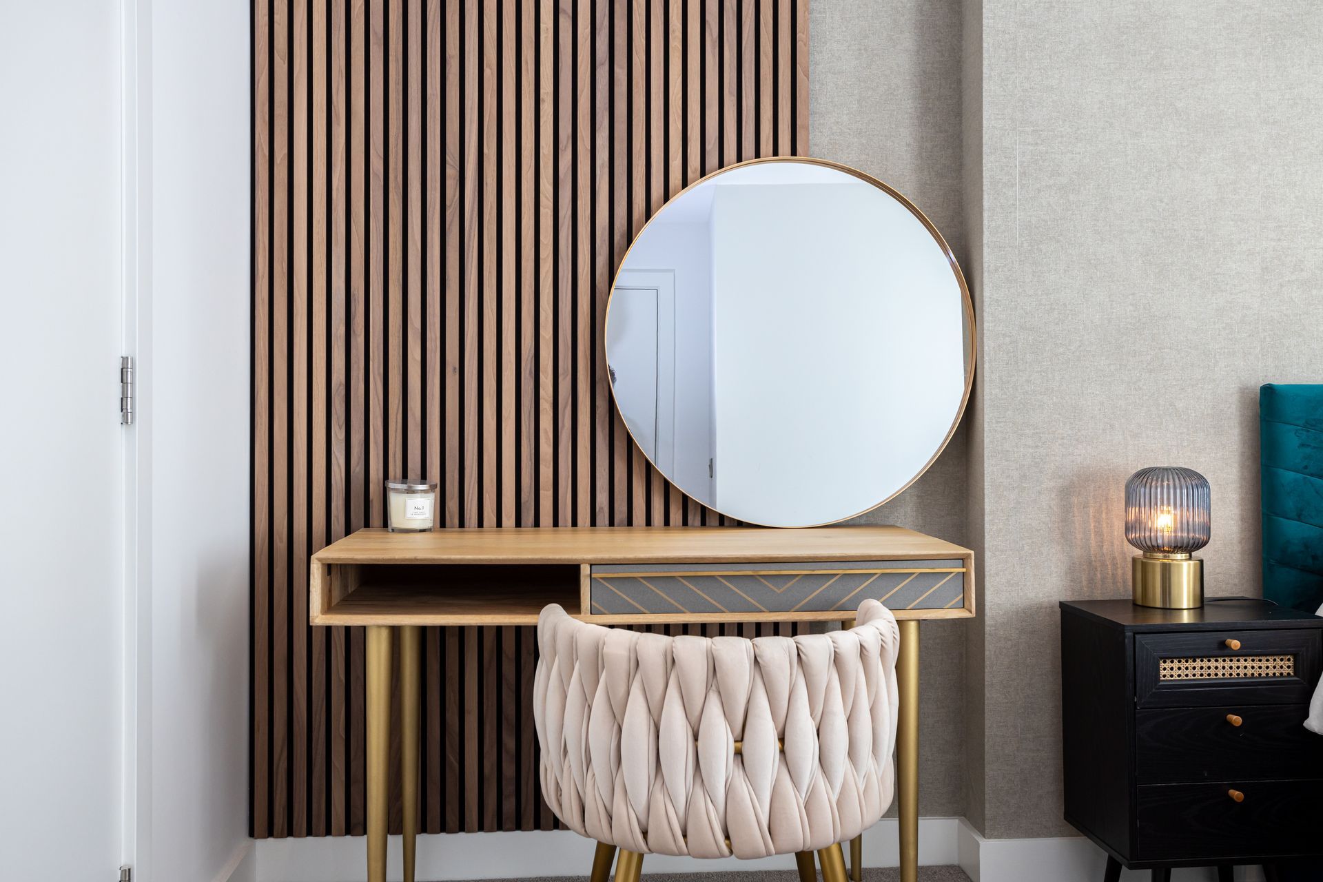 Vanity with round mirror, beige chair, wooden slat wall, and nightstand with lamp.