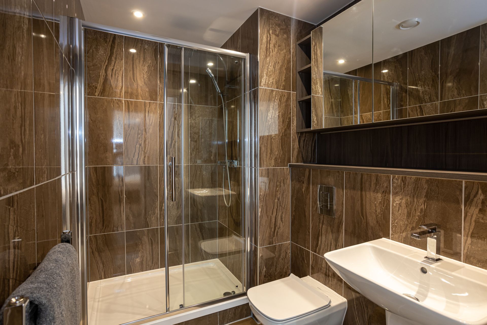 Brown-tiled modern bathroom with a glass shower, toilet, pedestal sink, and mirrored cabinet.