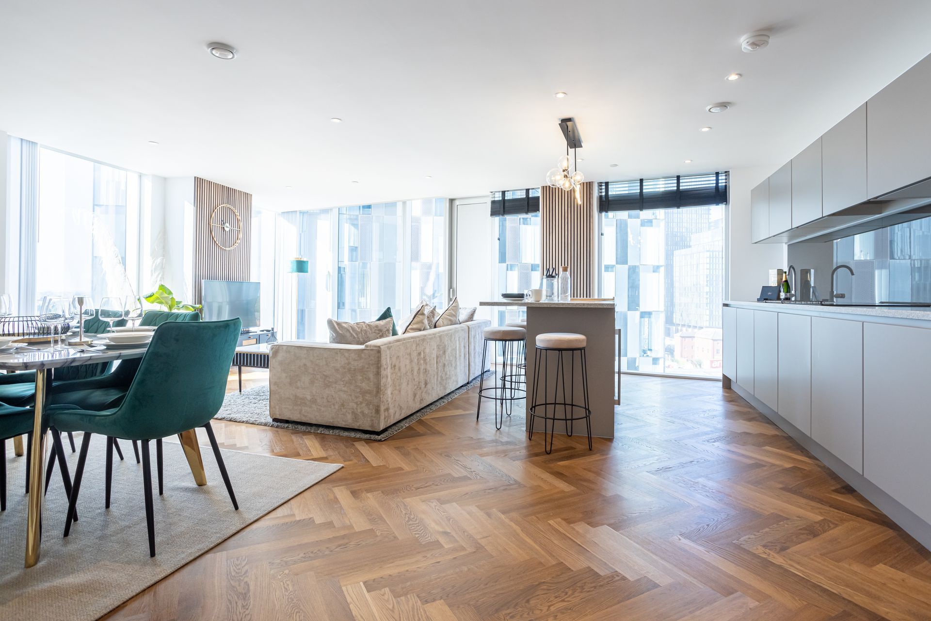 Modern open-plan apartment with dining, living, and kitchen areas, featuring wood floors, teal chairs, and large windows.