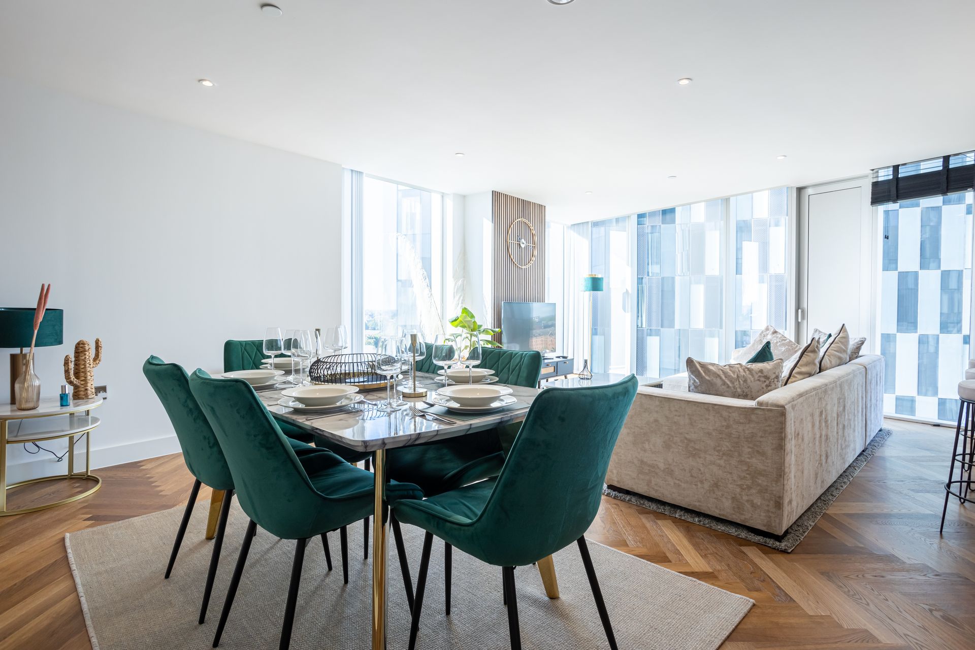 Dining area with marble table, emerald chairs, and a neutral-toned sofa. Natural light, wood floor.