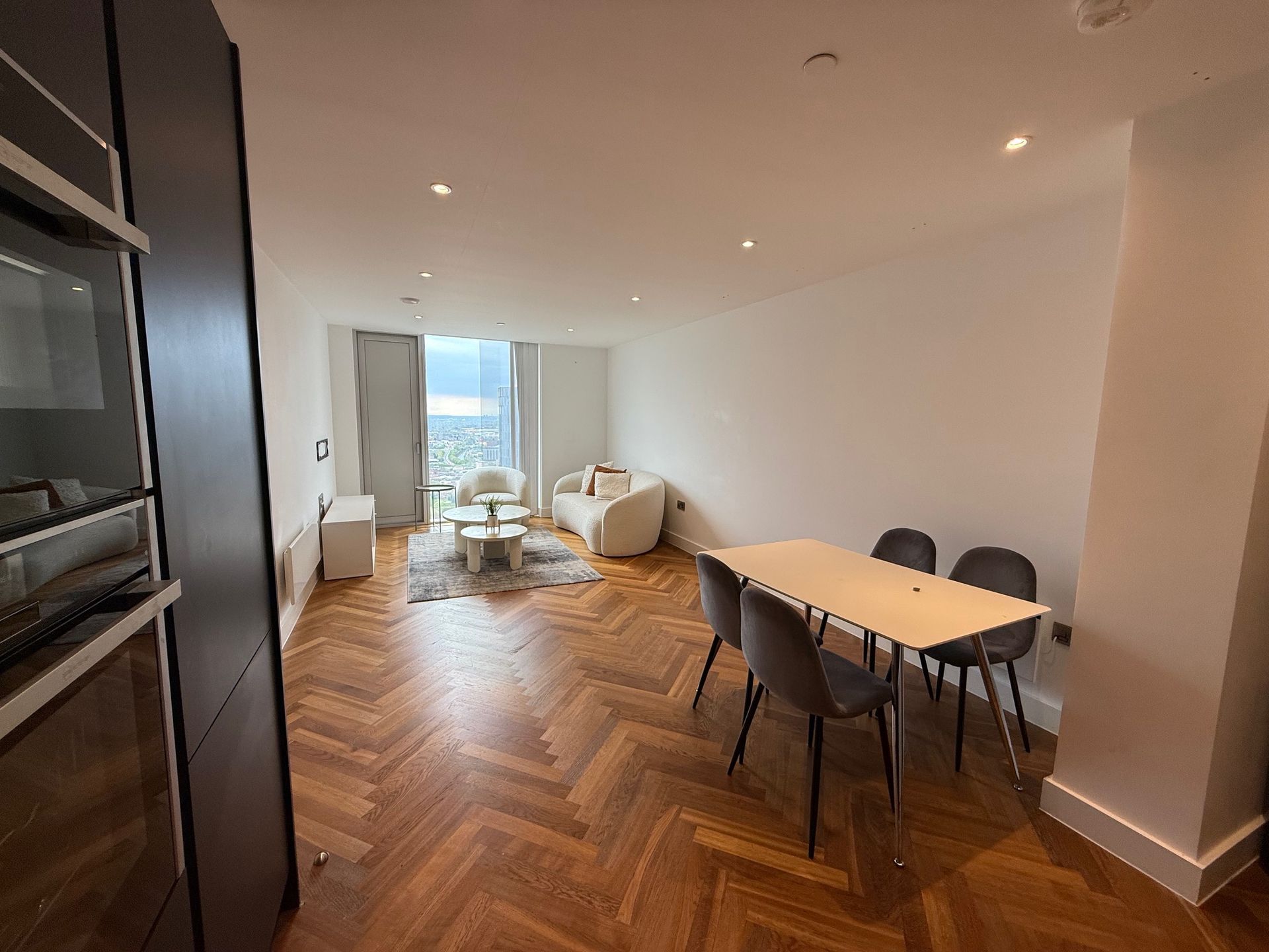 Living room with dining area: wooden floors, white walls, sofa, table, chairs, and large window view.