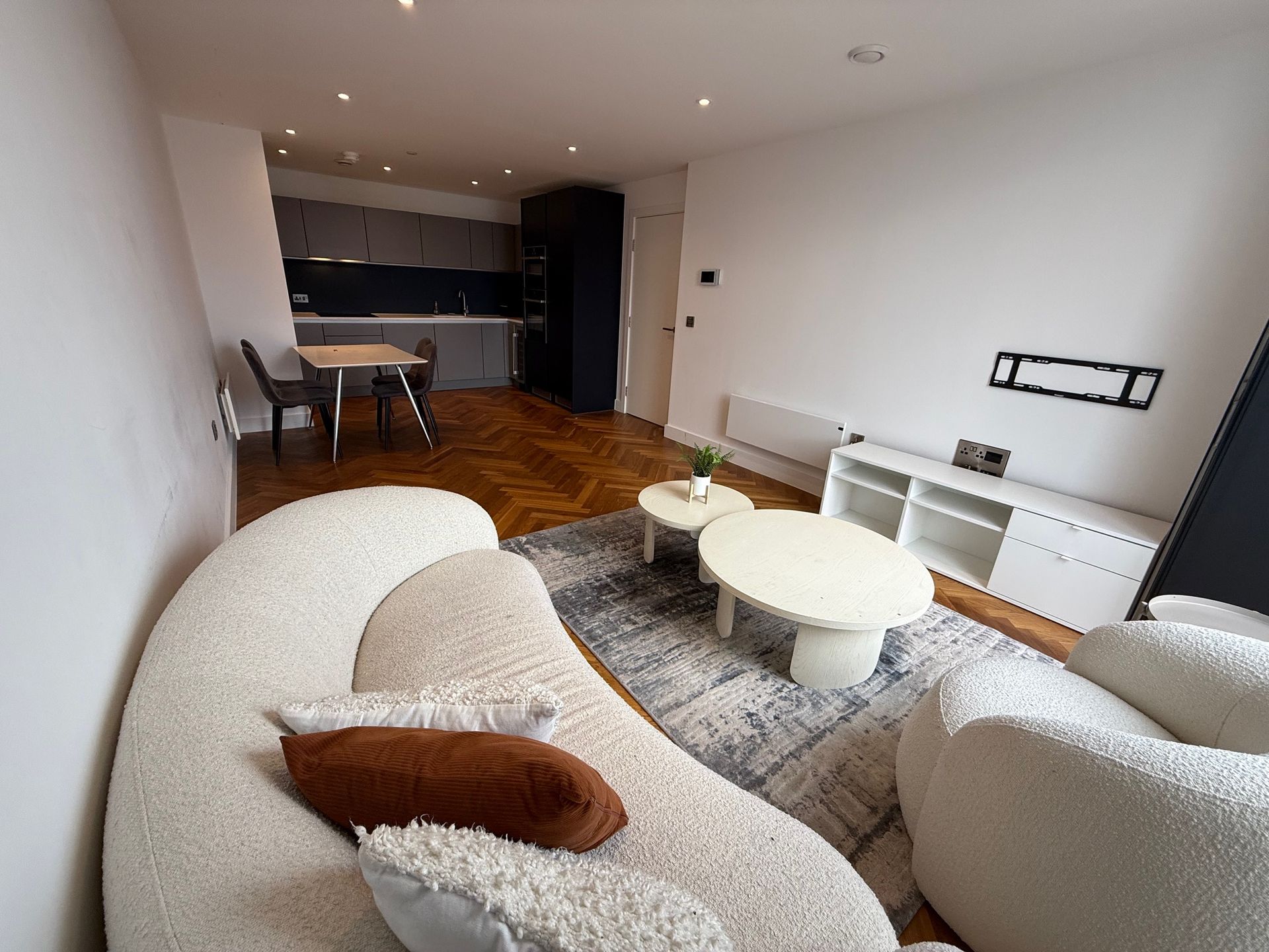 Living room with curved white sofa, coffee tables, TV stand, and open kitchen in the background.