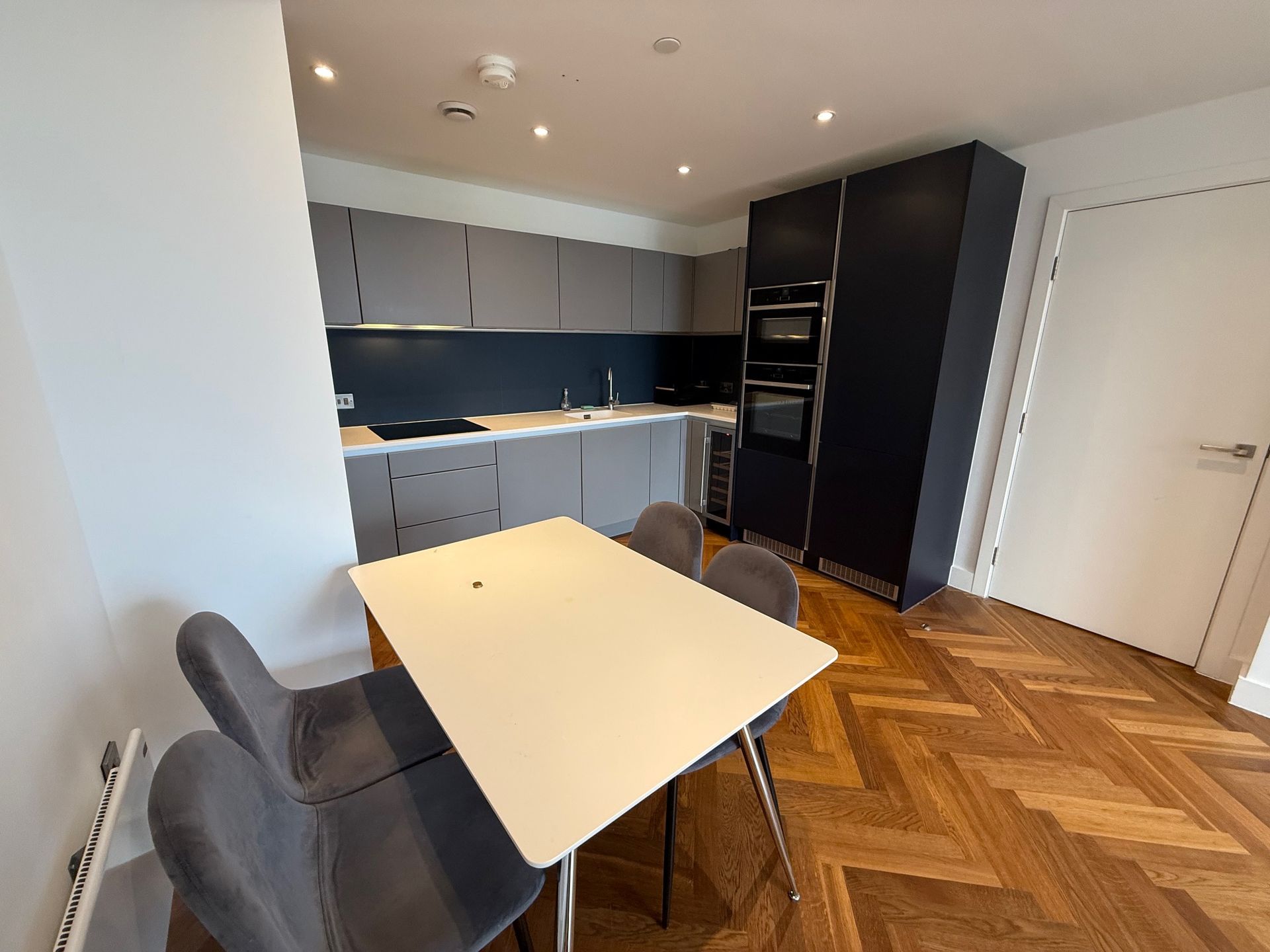 Modern kitchen with dining table, grey cabinets, and dark blue accent cabinets. Herringbone wood floors.