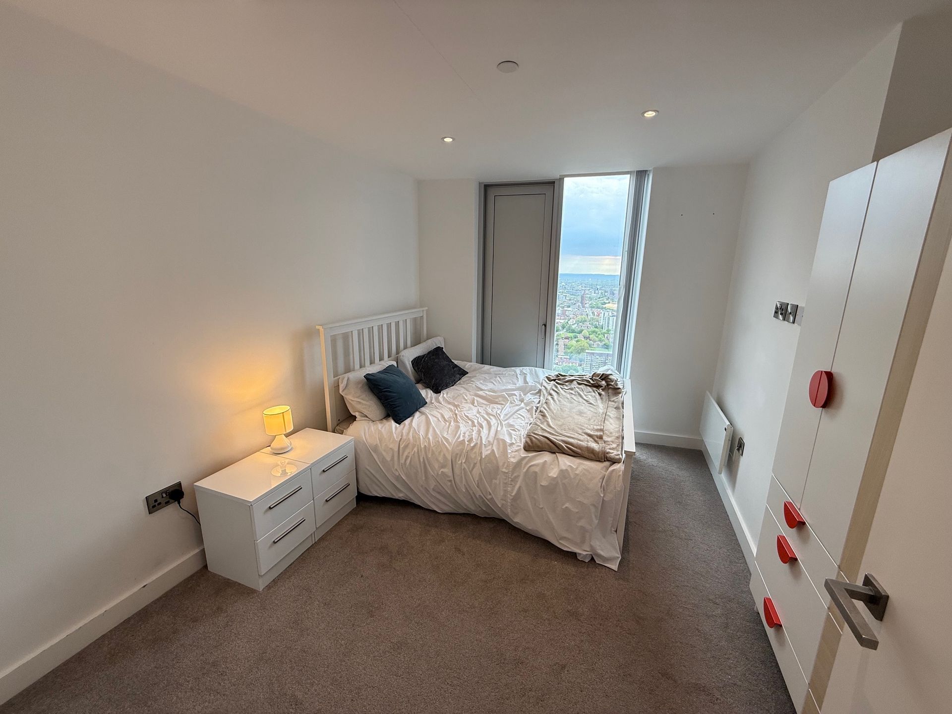 Bedroom with bed, nightstand, closet, window, and door. White walls, beige carpet.