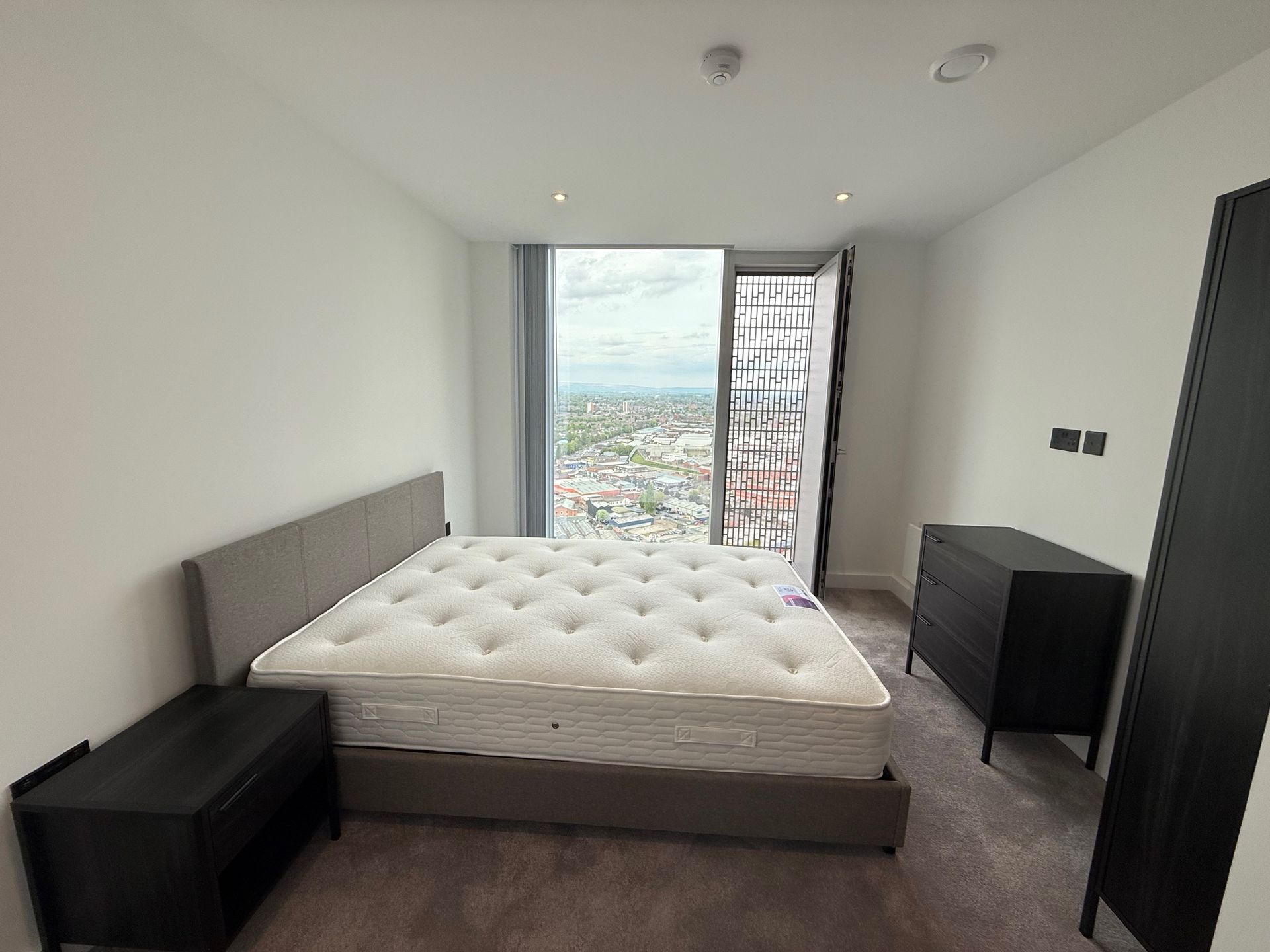 Bedroom with a large bed, dark furniture, and a cityscape view through a large window.