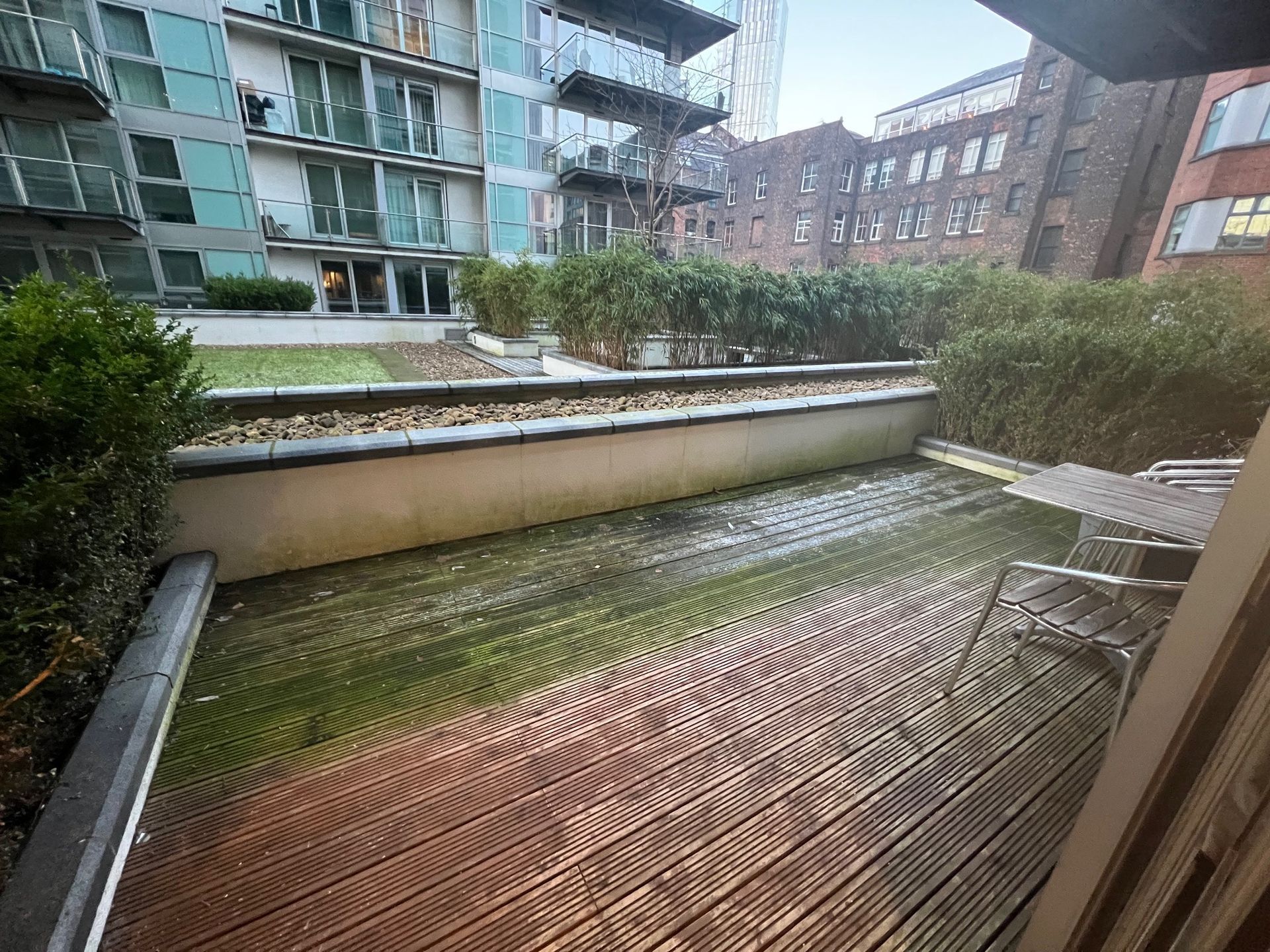 Wooden patio with a built-in planter featuring shrubs; apartment buildings in the background.