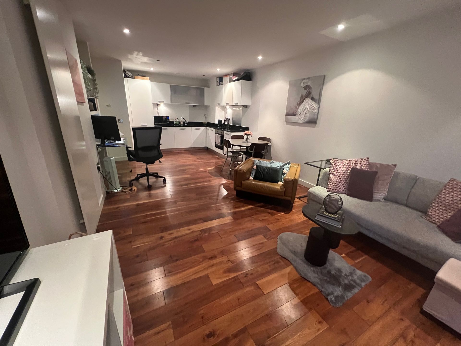 Living room with wooden floors, gray sofa, leather chair, and small kitchen in the background.