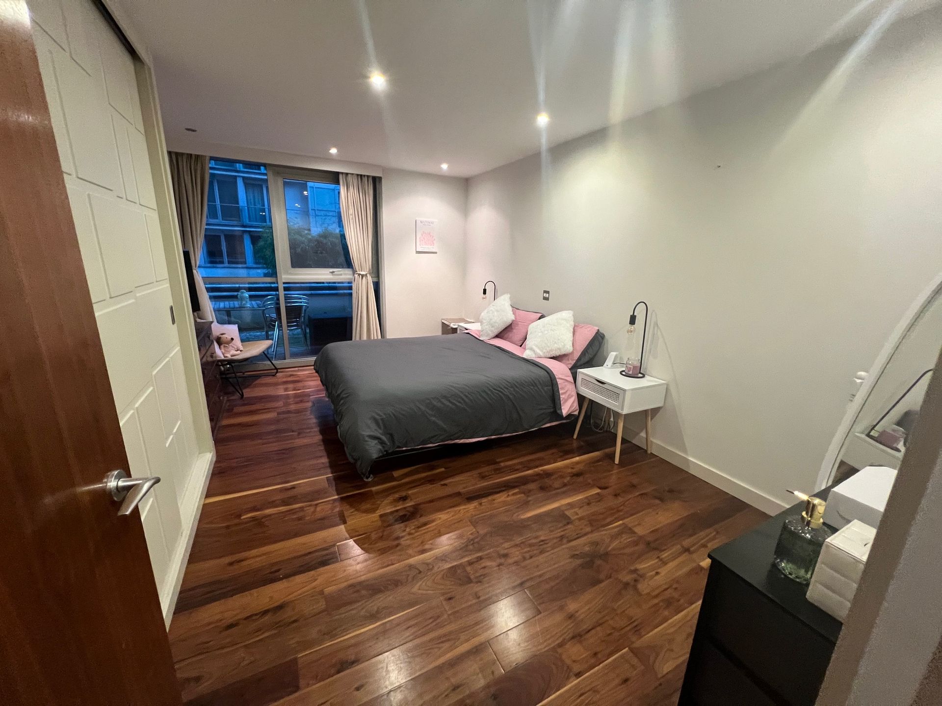 Bedroom with wooden floor, bed with gray comforter, white nightstand, mirror, and a door.