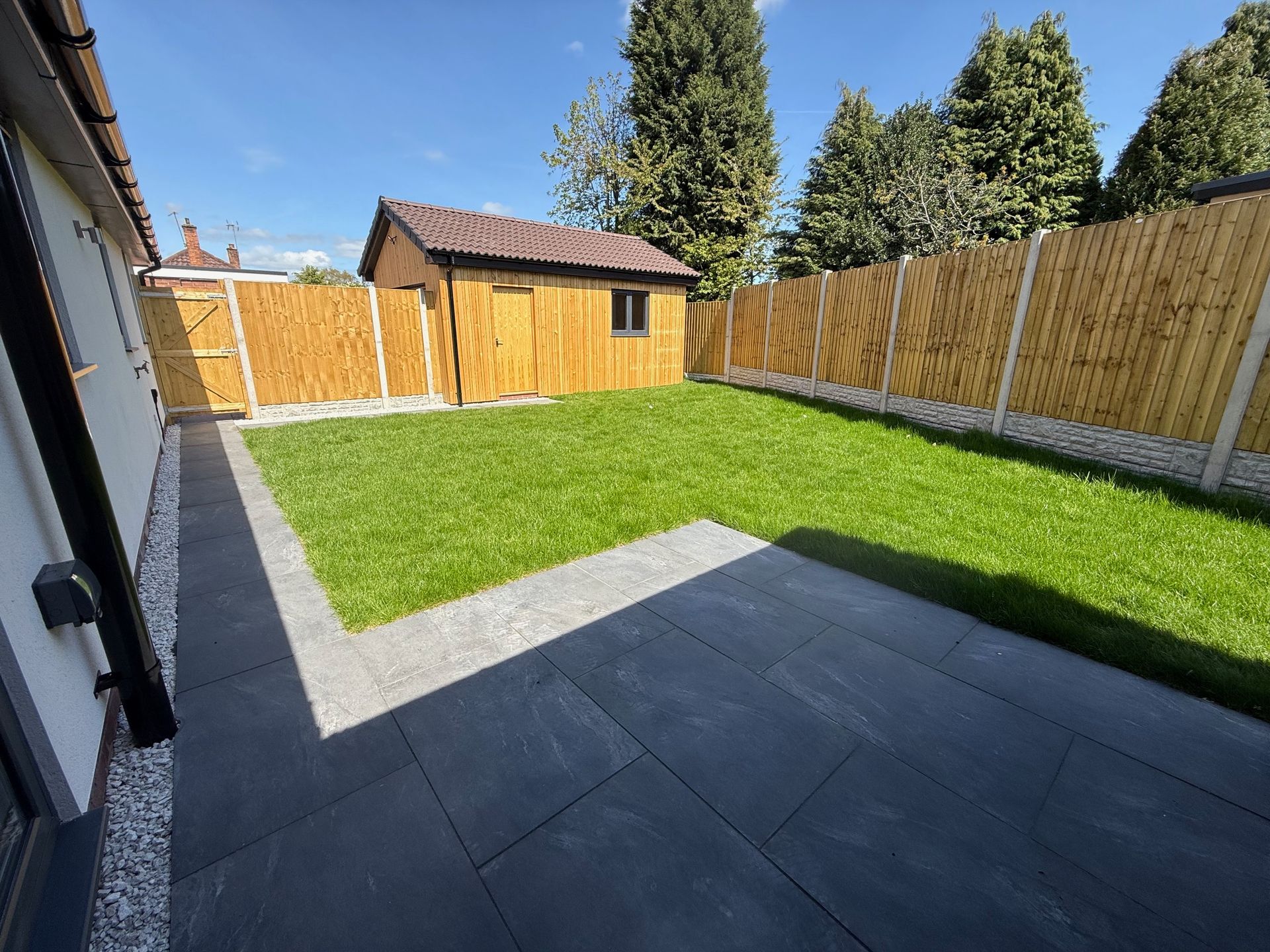 A backyard with a stone patio, green grass, a wooden shed, and a bamboo fence under a blue sky.