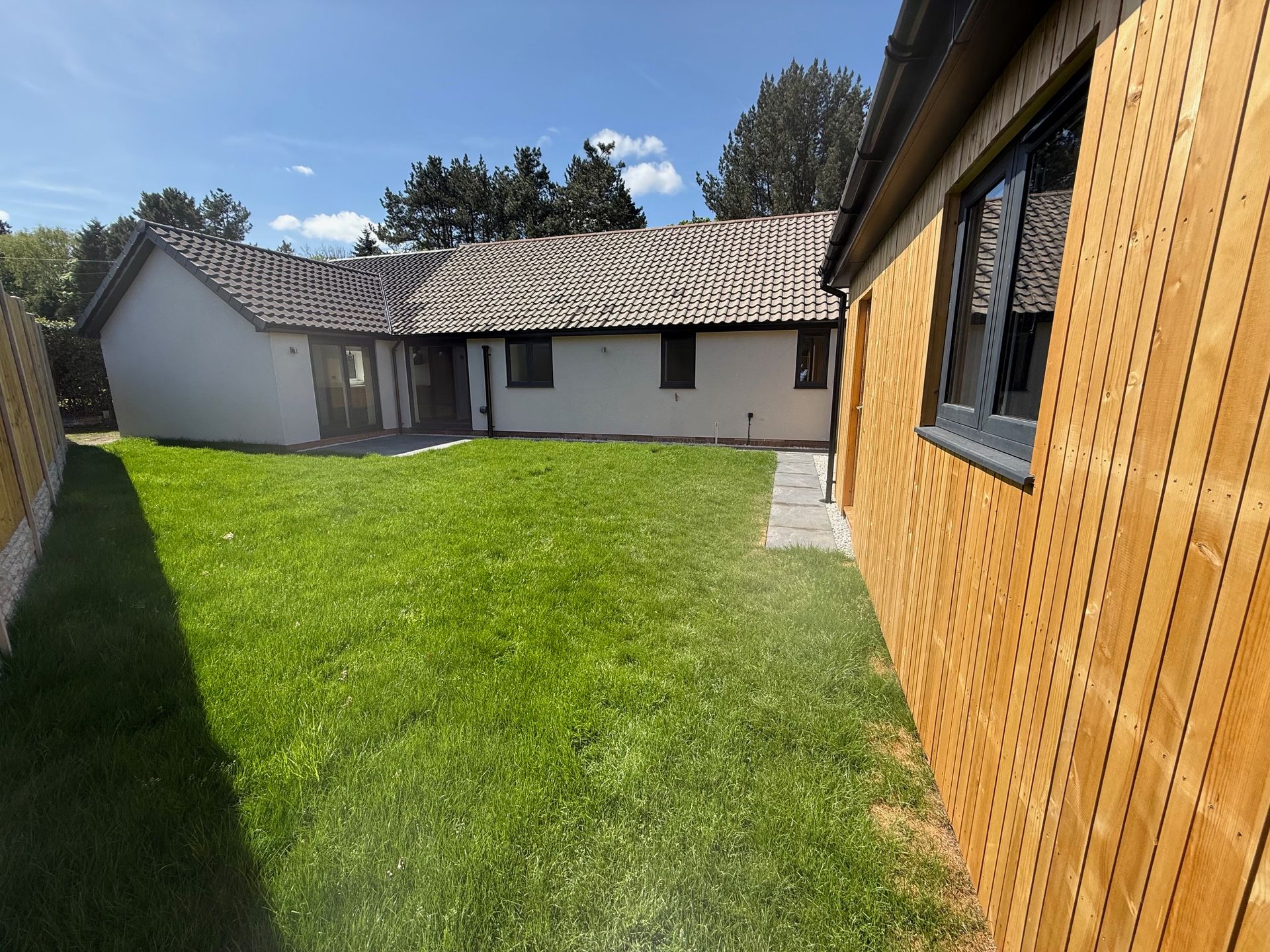A backyard with green grass, two buildings, and a wooden fence on a sunny day.