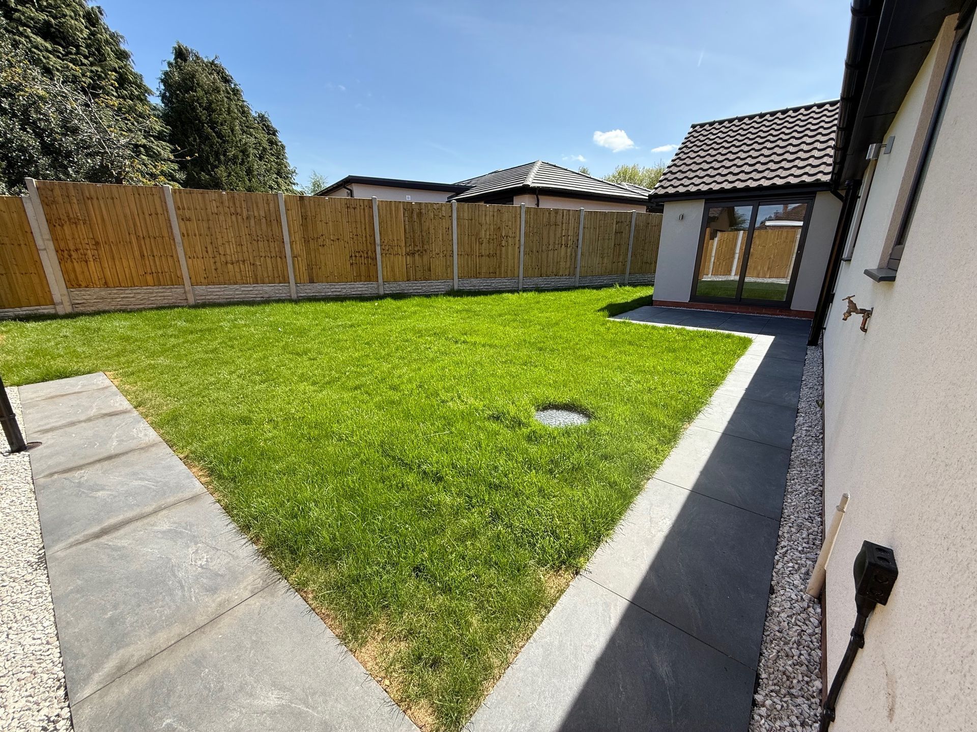 Lush green backyard with a grassy lawn, stone path, bamboo fence, and small building under a blue sky.