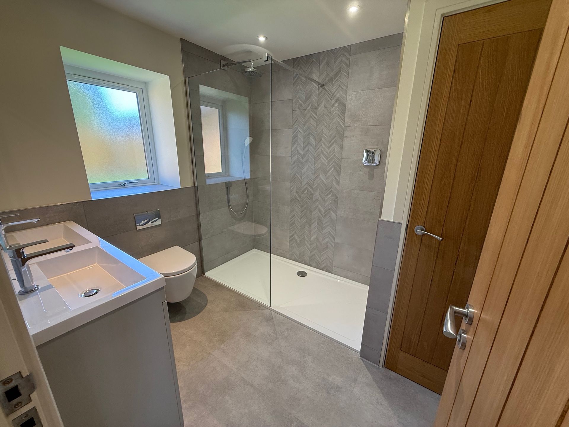 Modern bathroom with gray tiled walls and floor, shower, toilet, sink, and wooden door.