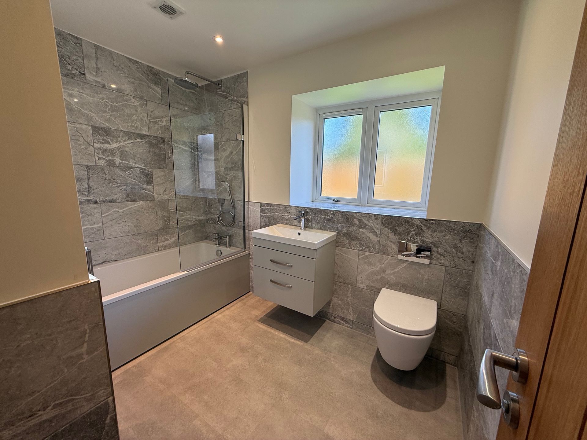 Modern bathroom with gray tiled walls and flooring, a white tub, sink, and toilet, and a window.