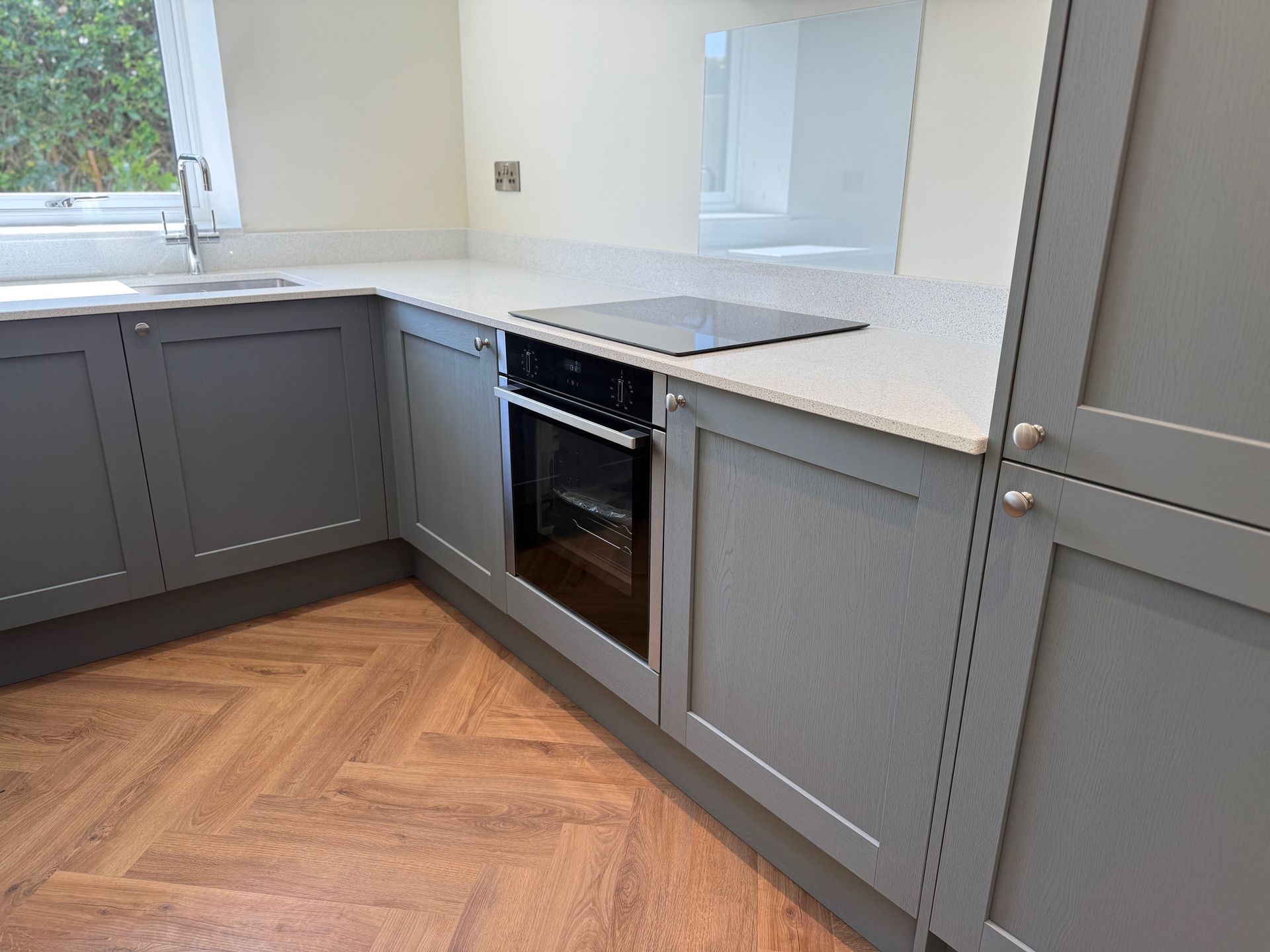 Gray kitchen cabinets and countertops with built-in oven and stovetop, herringbone wood-look floor.