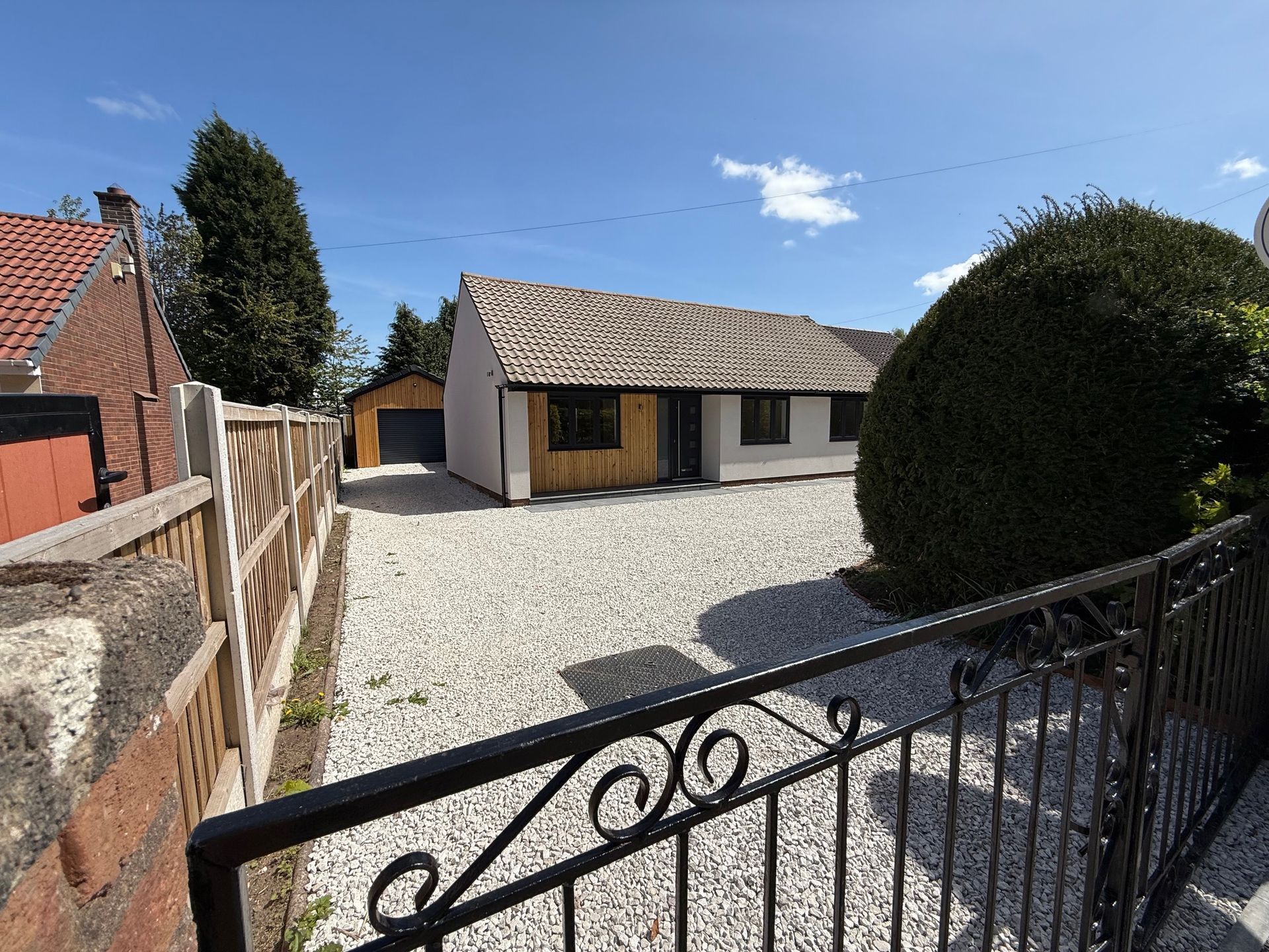 Bungalow with gravel driveway, garage, and front garden behind a black wrought iron fence under a blue sky.