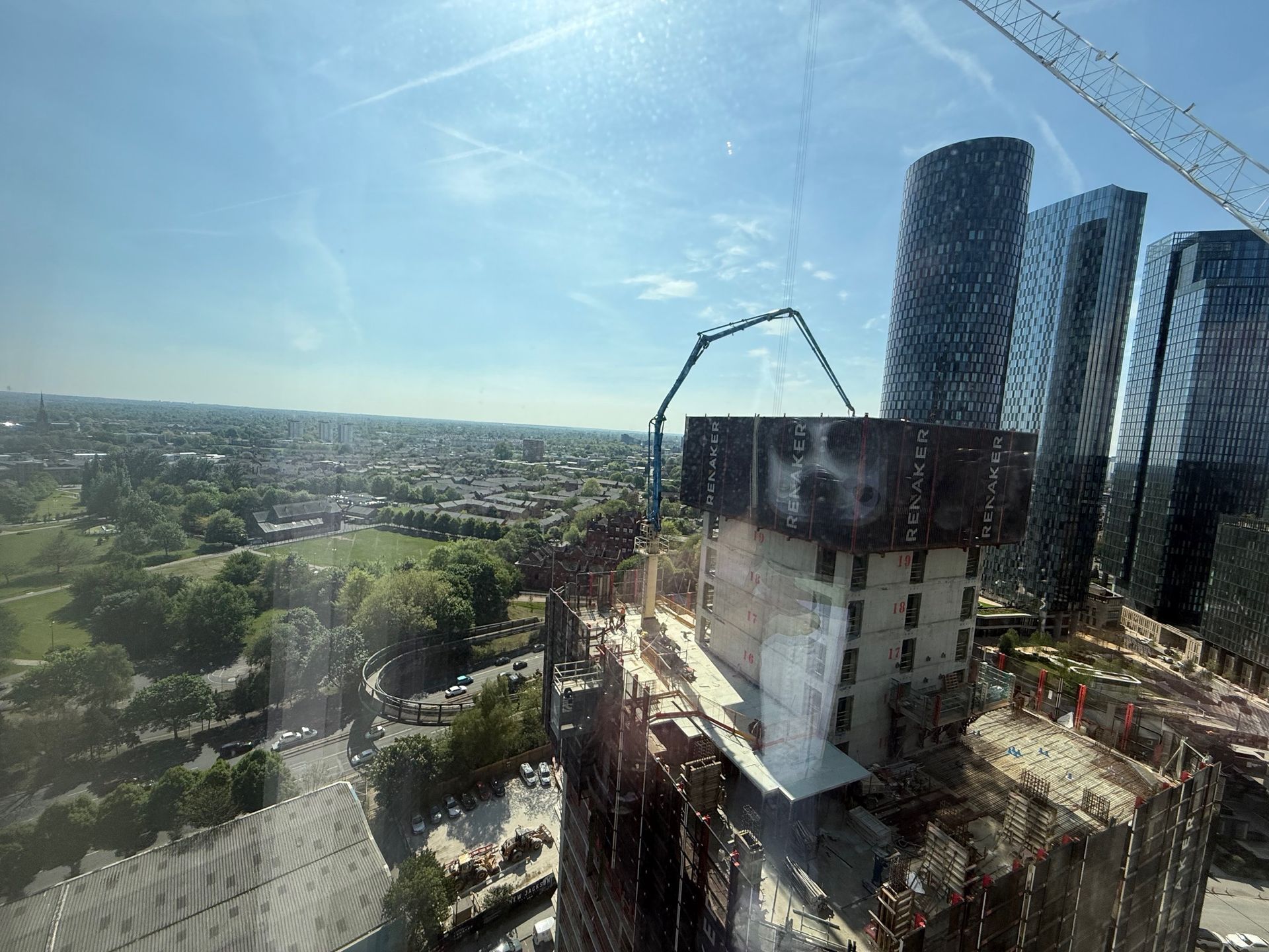 Construction site with tall buildings, concrete structure, crane, and green space visible in the background on a sunny day.