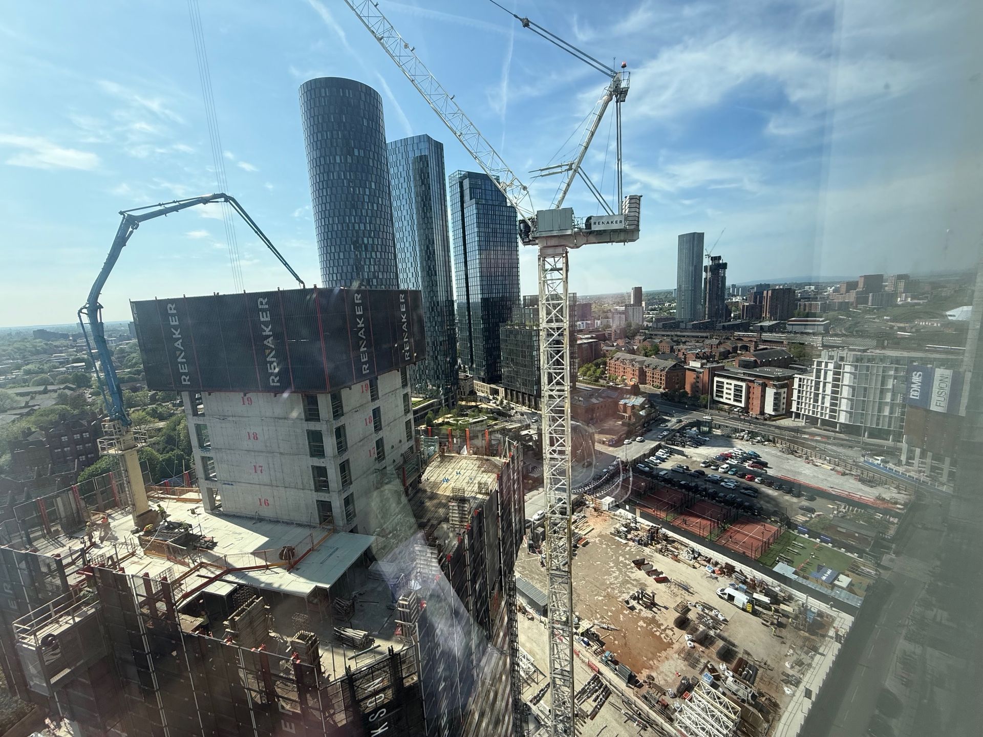 Construction site with tall buildings, cranes, and city skyline under a blue sky.