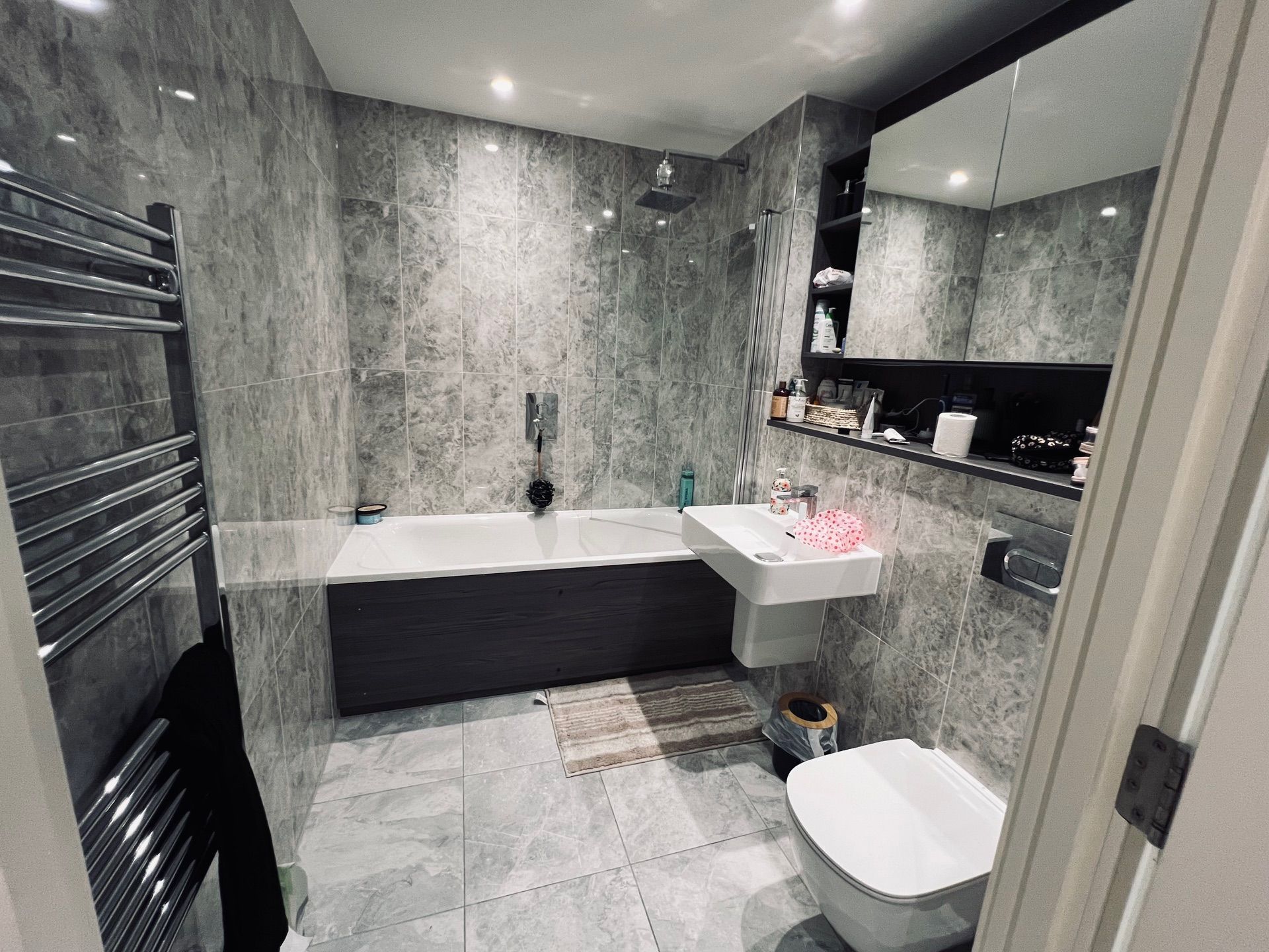 Modern bathroom with grey tile walls, bathtub, sink, toilet, and towel rack.