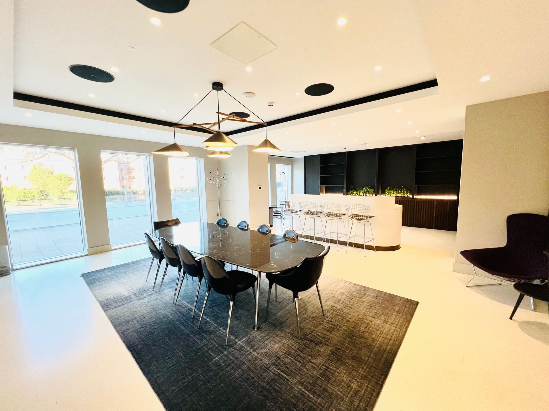 Spacious dining area with long table, black chairs, and modern lighting. Bright room, rug, and bar in the background.