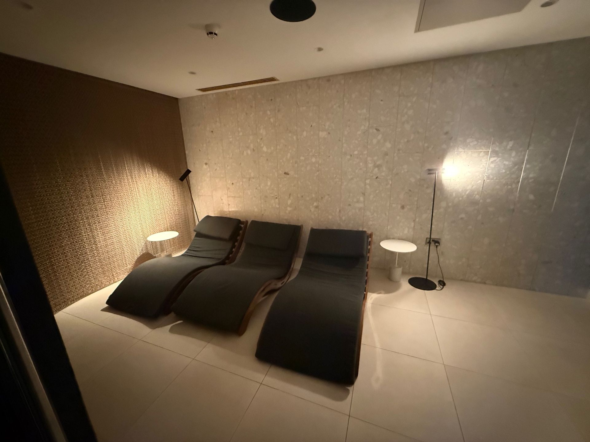 Three dark chaise lounge chairs in a relaxation room with mosaic tile walls and floor, two small side tables and lamps.
