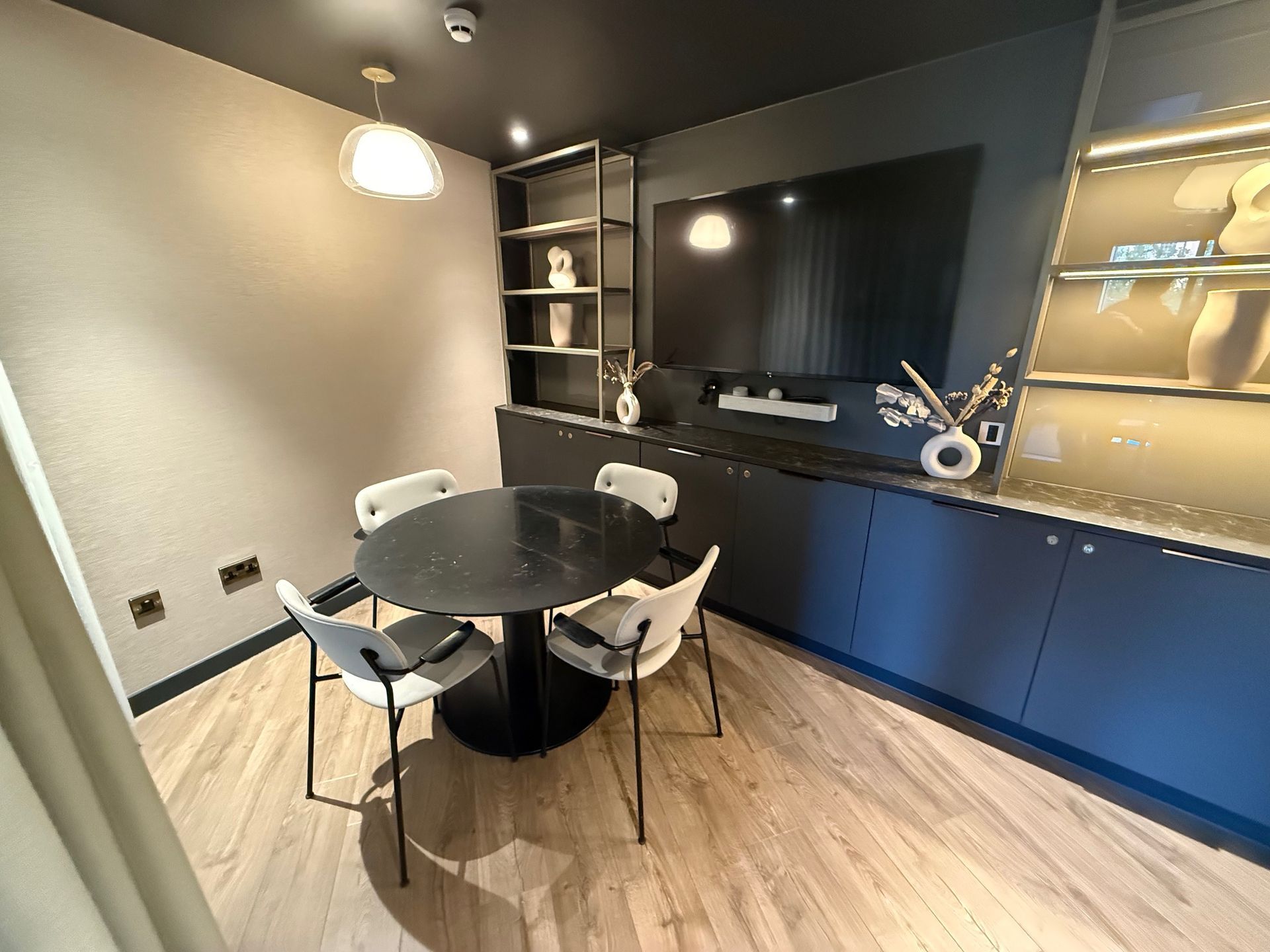 A small dining area with a round black table and four white chairs. Includes built-in cabinets and a TV.