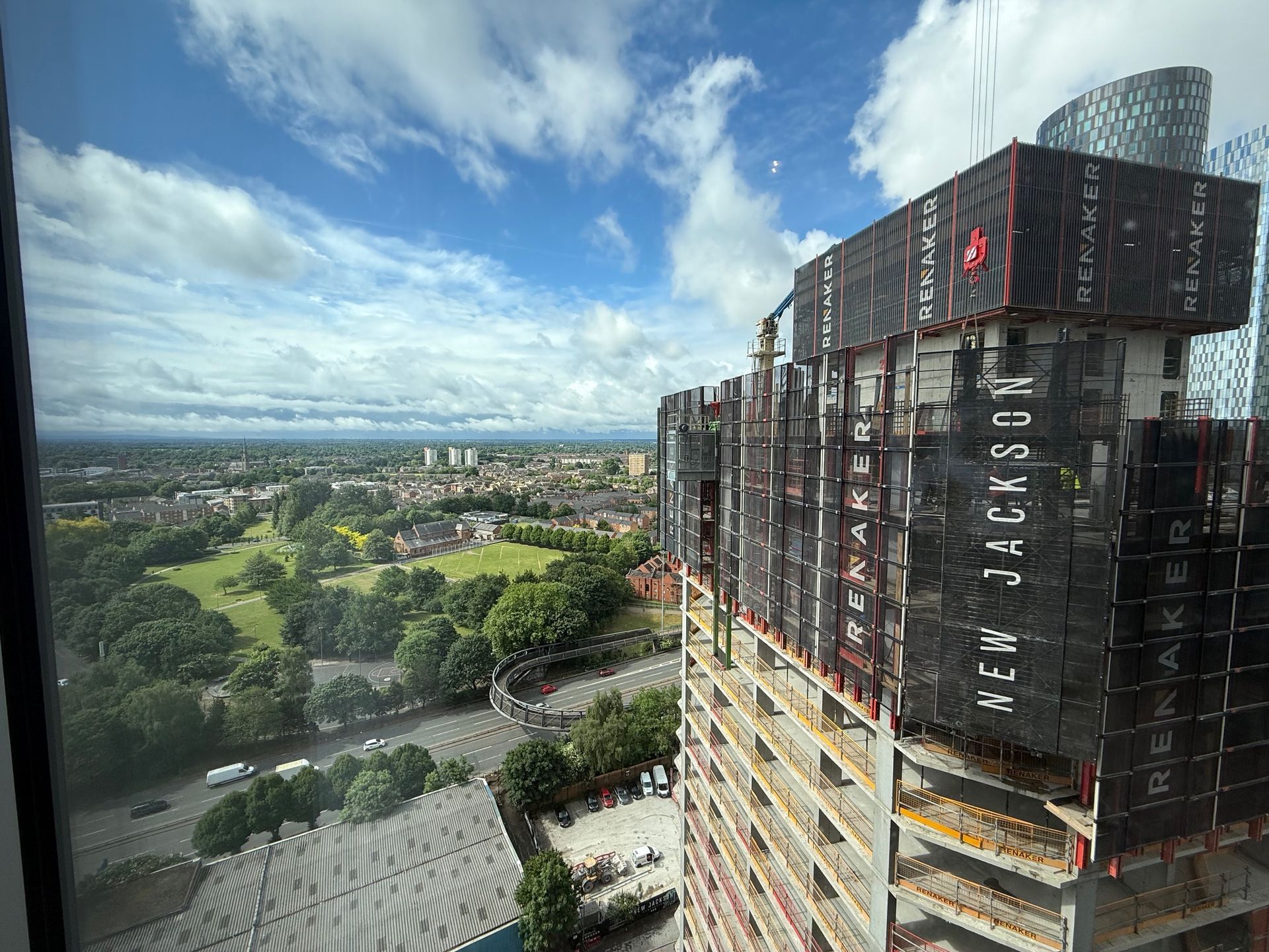 High-rise construction site with a partially built skyscraper against a backdrop of a city and green park under a cloudy sky.