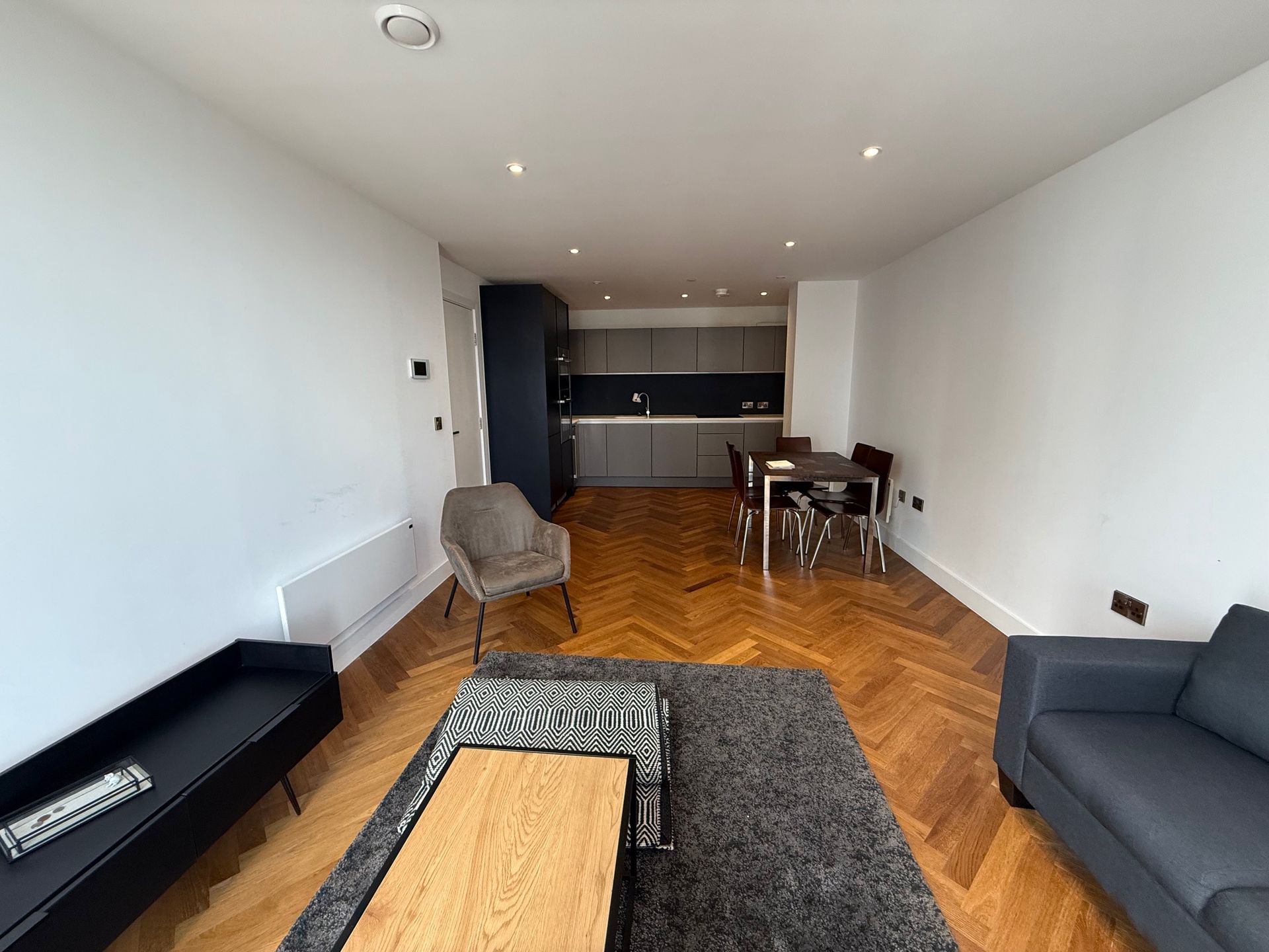 Living room with wood floors, gray furniture, and a kitchen in the background.