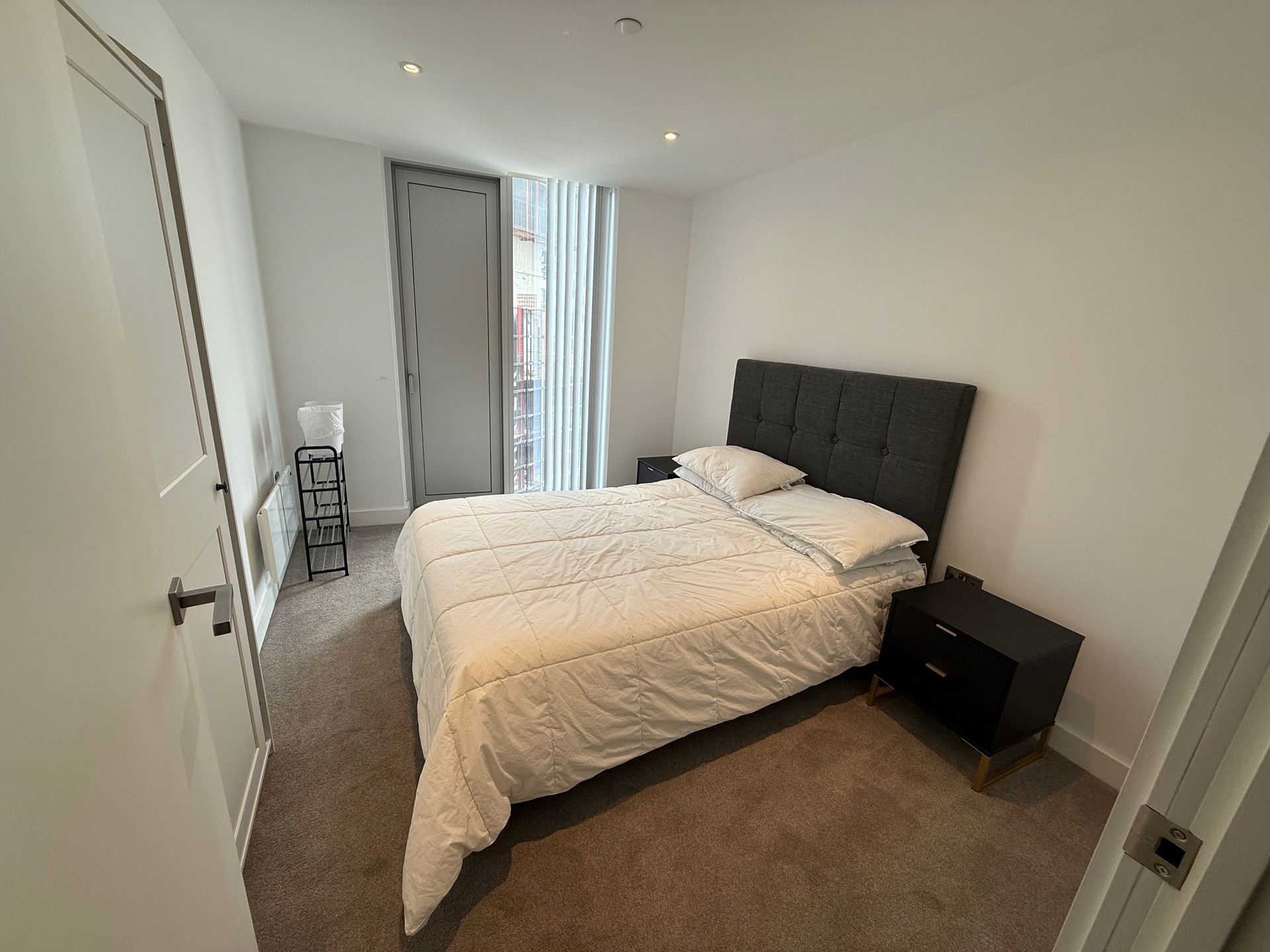 Bedroom with a bed, nightstand, door, and a radiator. Bright, white walls, and a window with blinds.