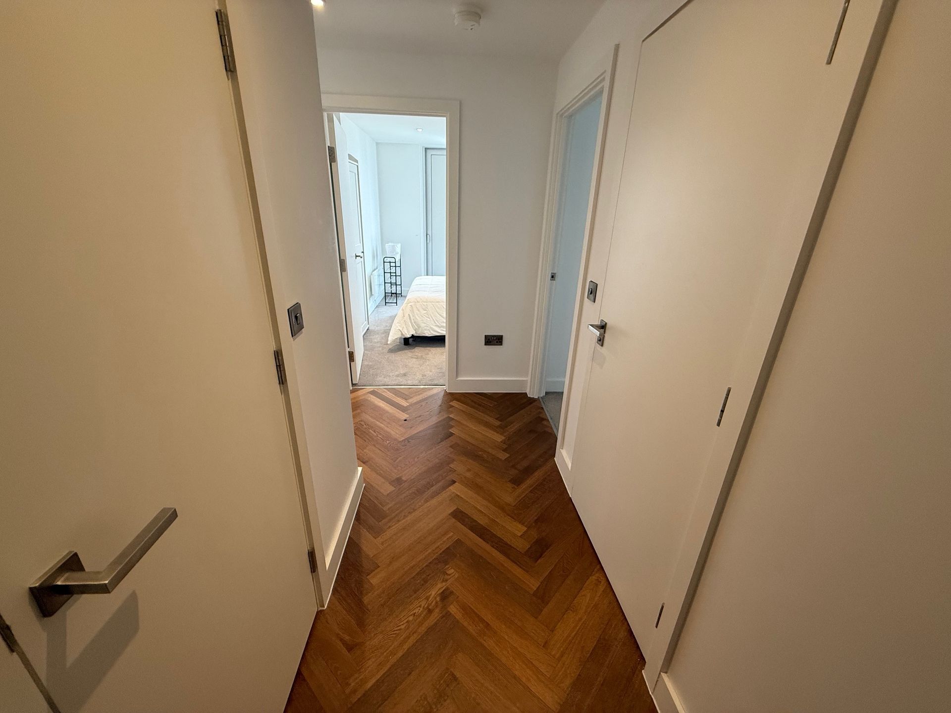Narrow hallway with herringbone wood floor and white doors leading to a bedroom.