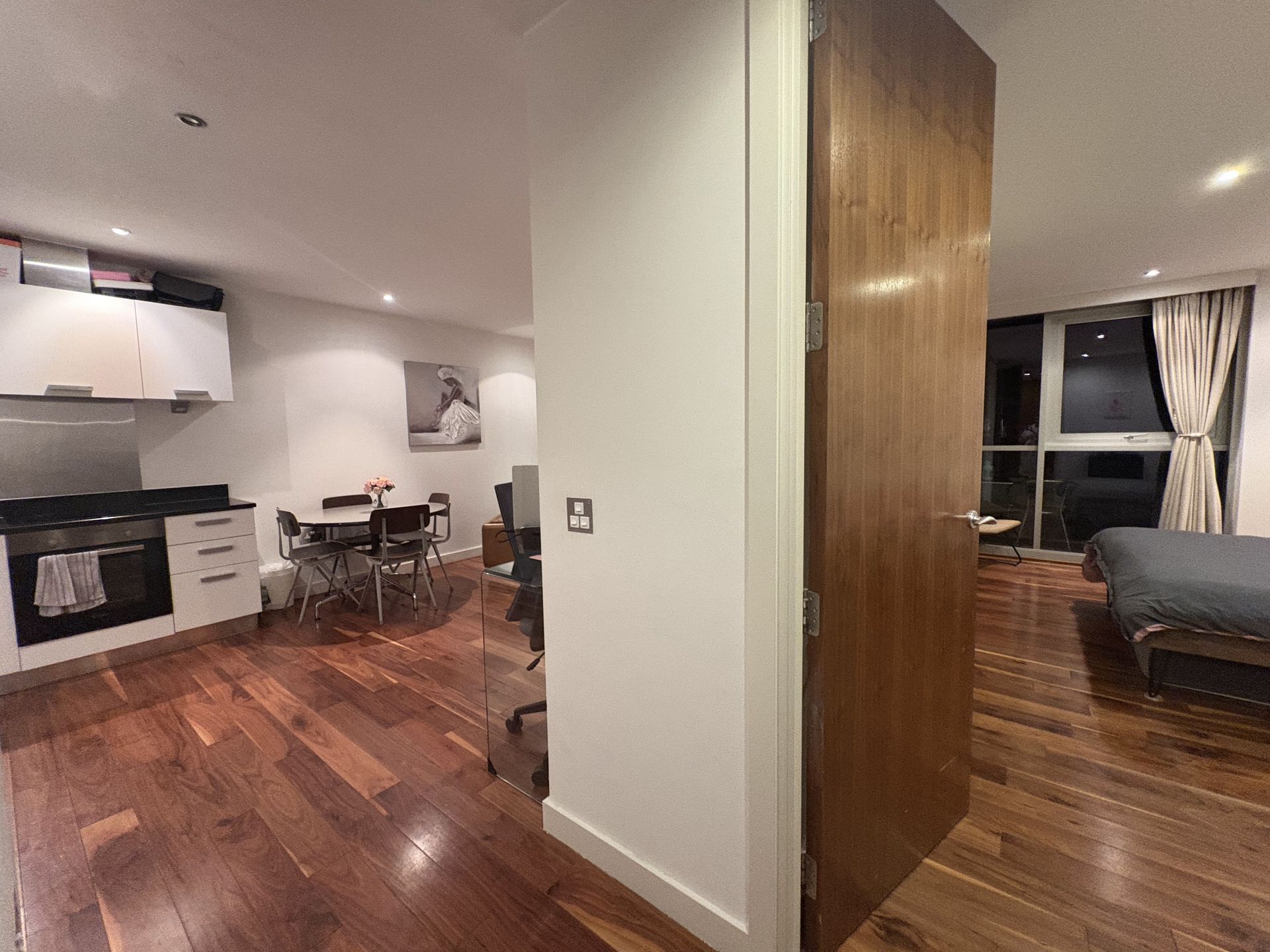 Interior view of an apartment with a kitchen, dining area, and bedroom, featuring wooden floors and dark wood door.