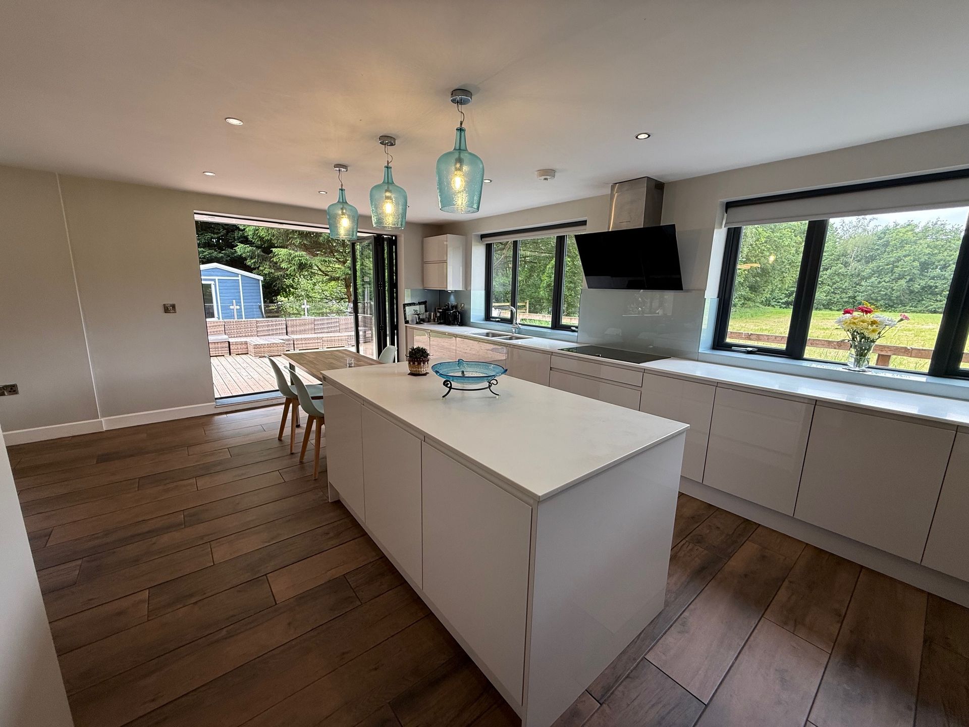 Modern white kitchen with island, wooden floors, and glass pendant lights. Sliding doors open to a deck.
