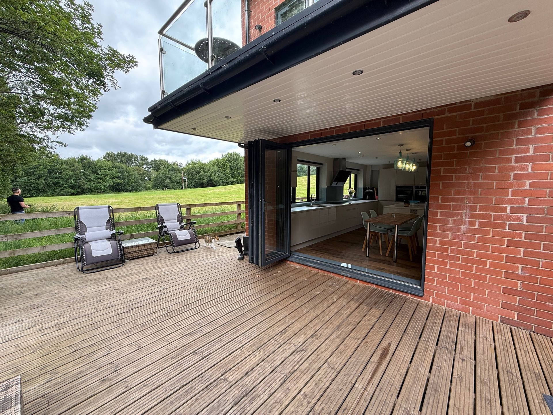 Wooden deck with open doors to a modern kitchen, two chairs, red brick wall, and a grassy field.