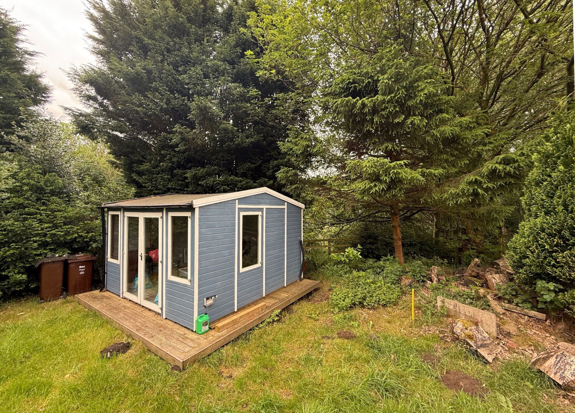 Blue shed with windows and wooden deck in a grassy, wooded area.