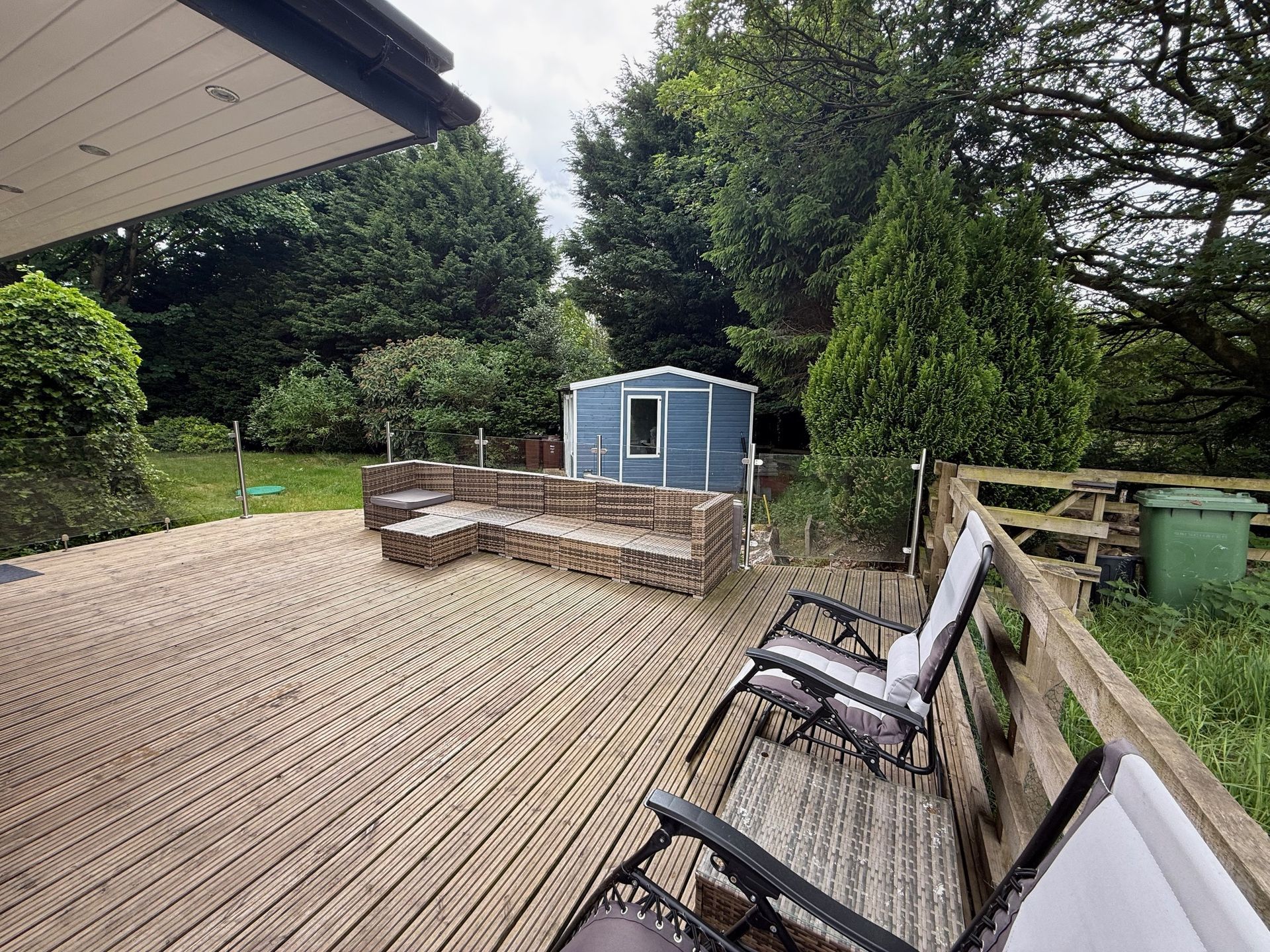 Wooden deck with seating and a shed in a backyard with trees.