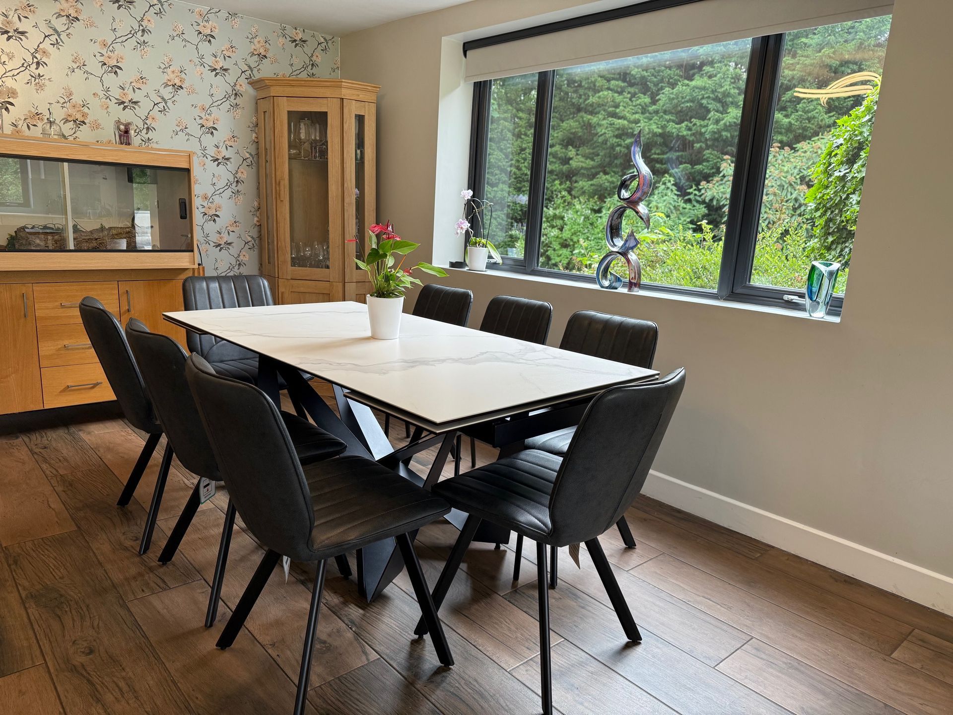 Dining room: white marble table, eight gray chairs, wood floor, floral wallpaper, large window with greenery view.