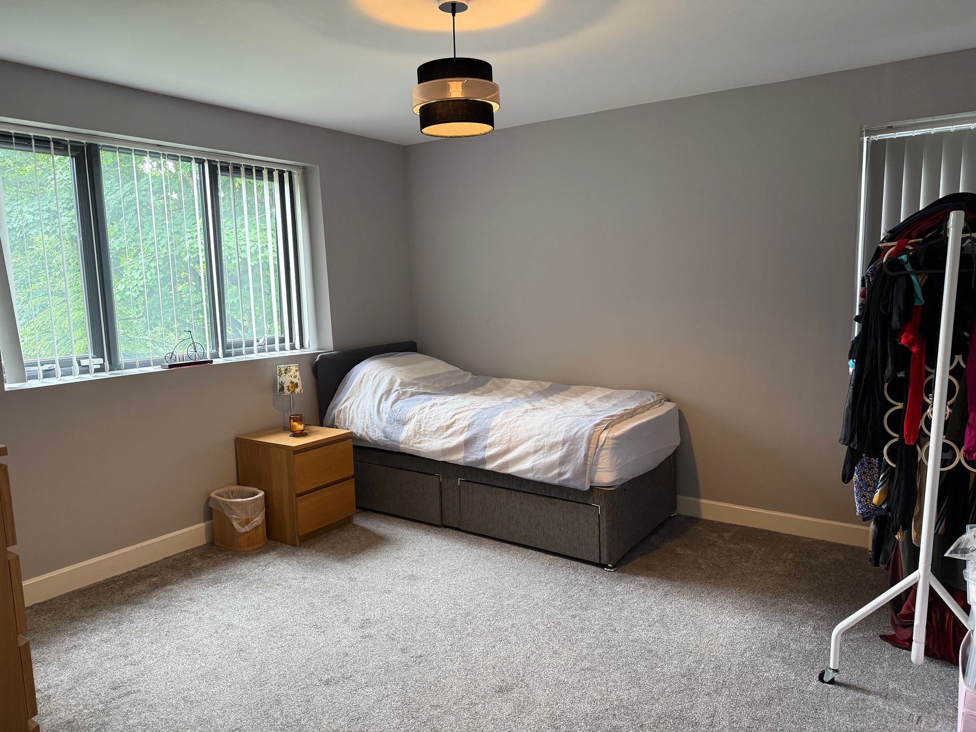 Bedroom with bed, nightstand, window, clothing rack, and gray walls.