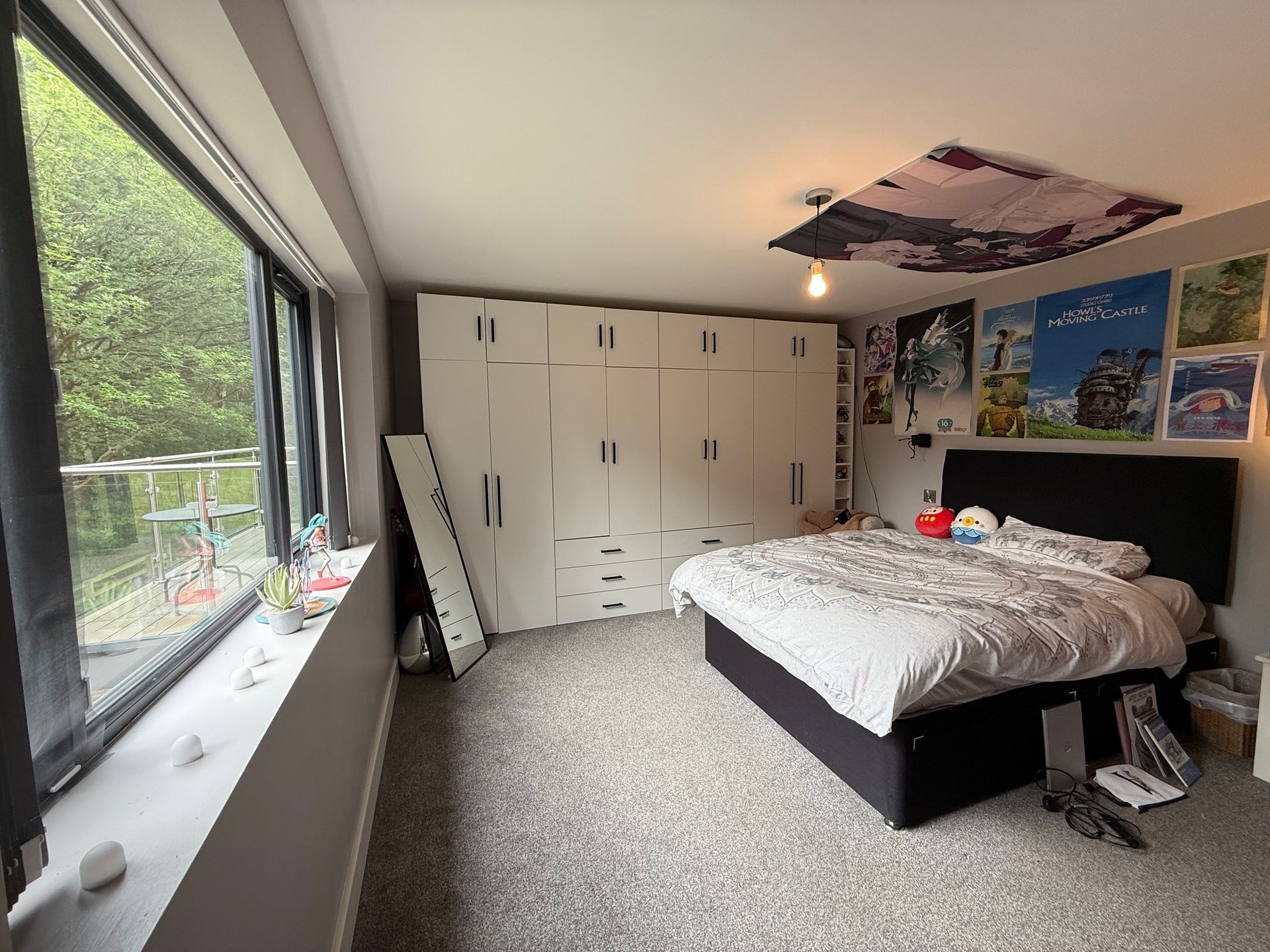 Bedroom with large window, white wardrobe, bed, posters on the wall, and gray carpet.