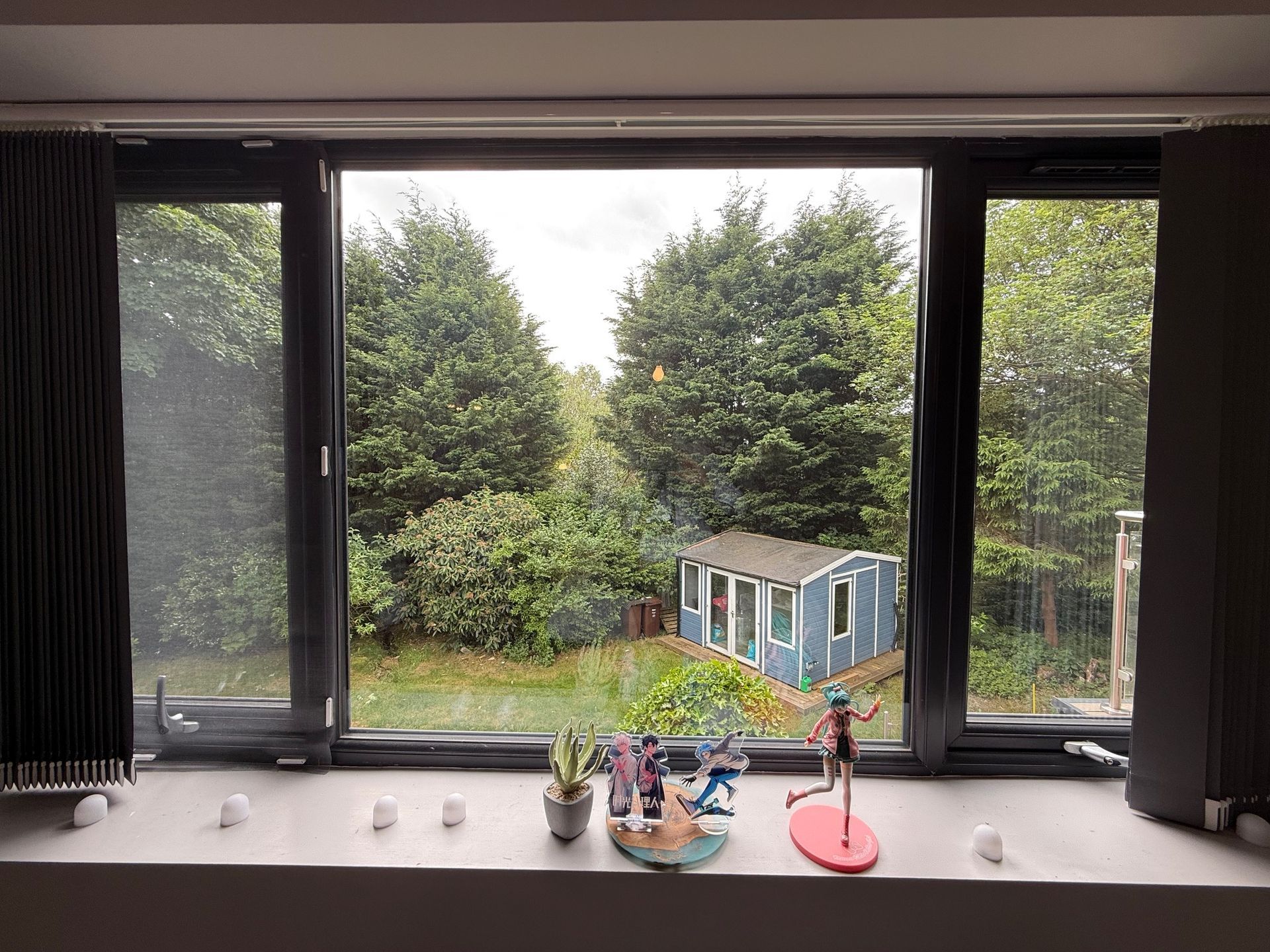 View through a window of a backyard with trees and a blue shed. Window sill displays figurines and snowballs.