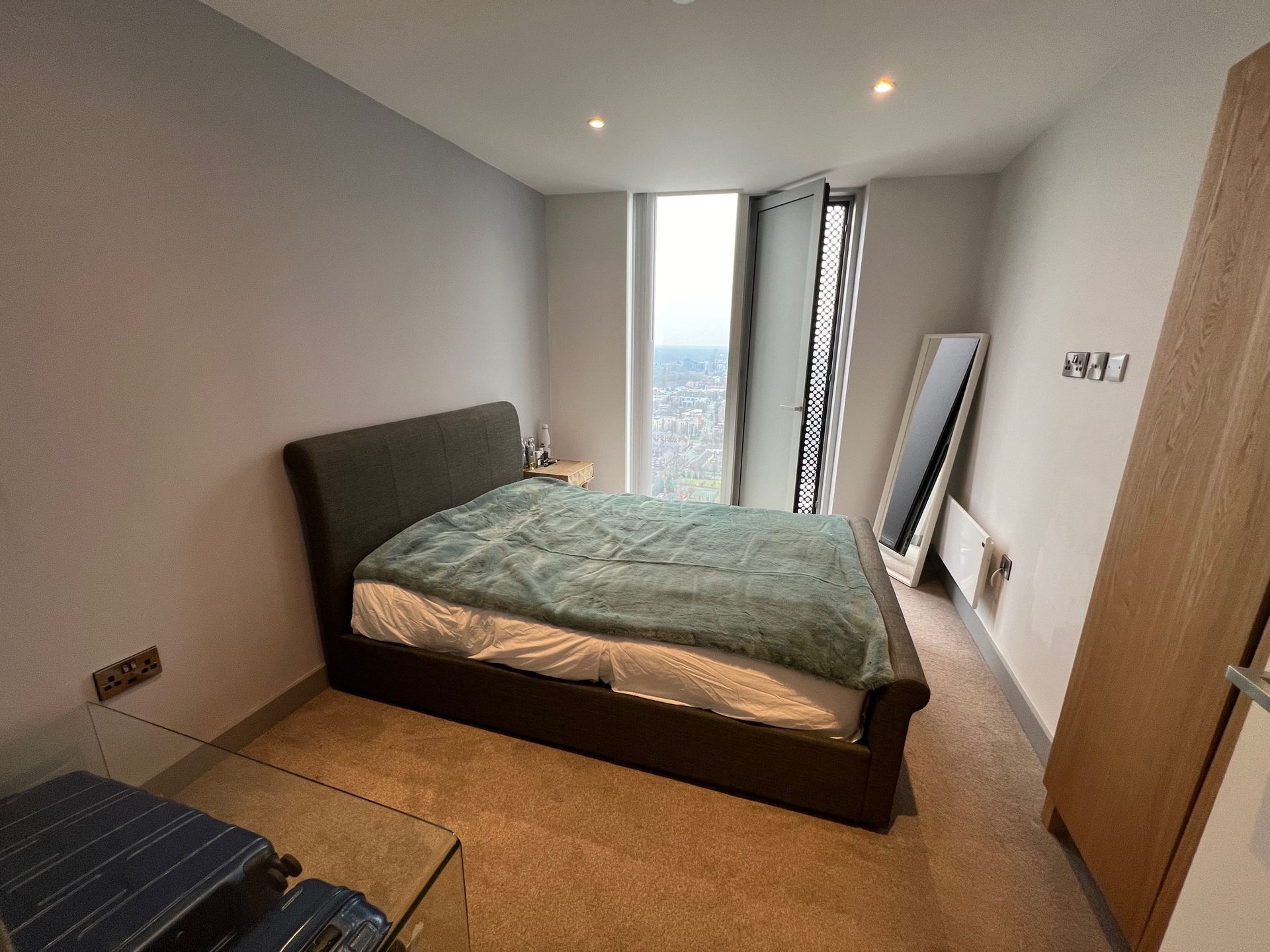 Bedroom with bed, tall window, mirror, and cabinet. Gray walls, brown bed frame, and tan carpet.