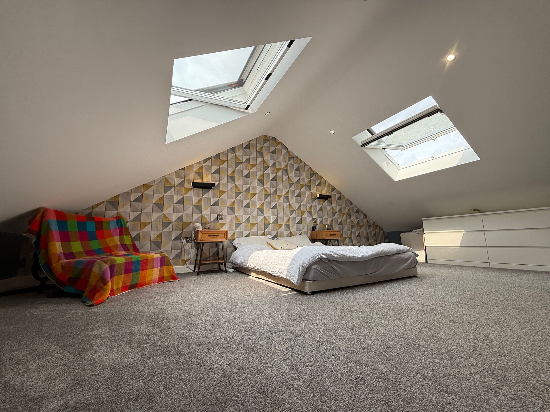 Bedroom with bed, sofa, geometric wallpaper, and skylights in a sloped ceiling. Carpeted floor.