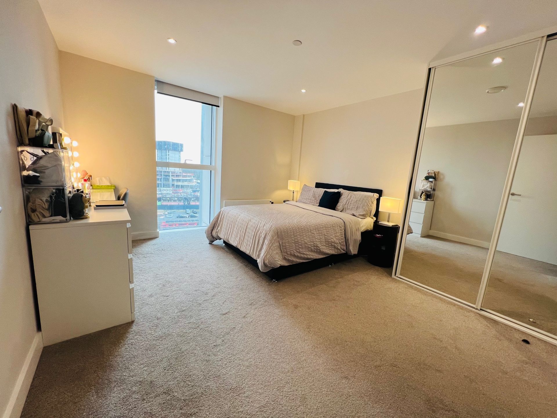 Bedroom with bed, desk, full-length mirror, and window with city view. Neutral tones, carpeted floor.